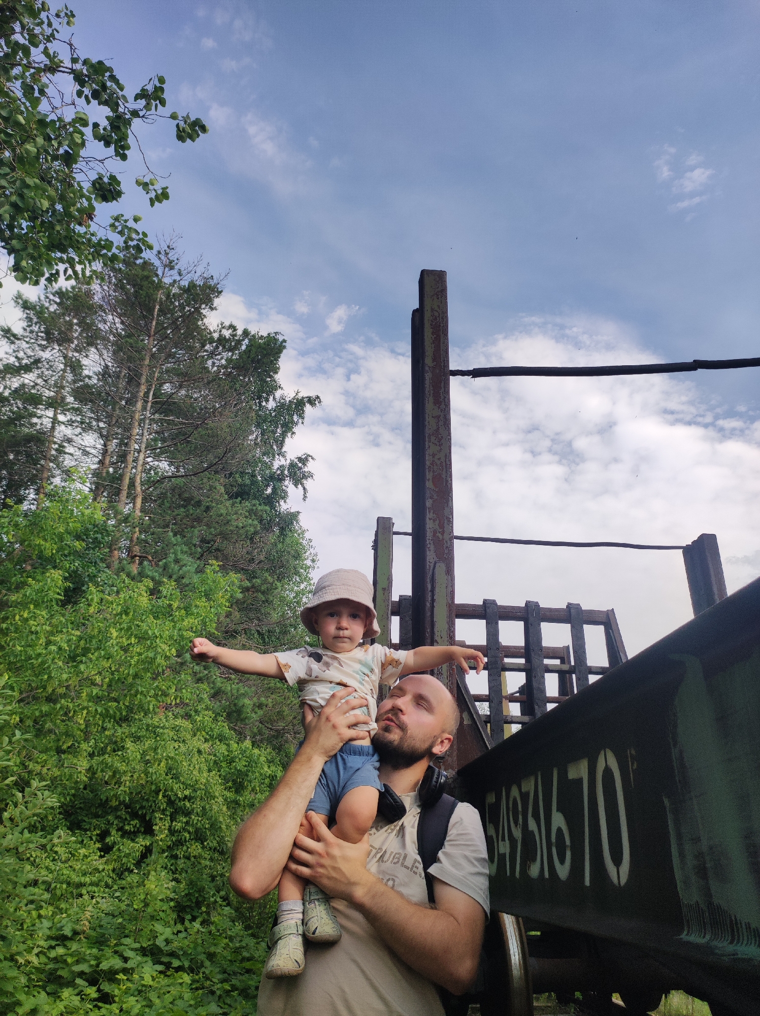 A bald man with his eyes closed is holding a young child on his shoulders. The man is wearing a light-colored t-shirt and a backpack. The child is wearing a light-colored hat, a patterned t-shirt, and blue shorts. The child's arms are outstretched. They are standing outdoors next to a dark green train car with the number "54931670" painted on its side. Behind them is a dense forest with tall trees. The sky is partly cloudy and blue. The scene is likely in an industrial area or a place with abandoned railway equipment, possibly on the outskirts of Chaikovsky, Russia, given the presence of such infrastructure near natural landscapes.