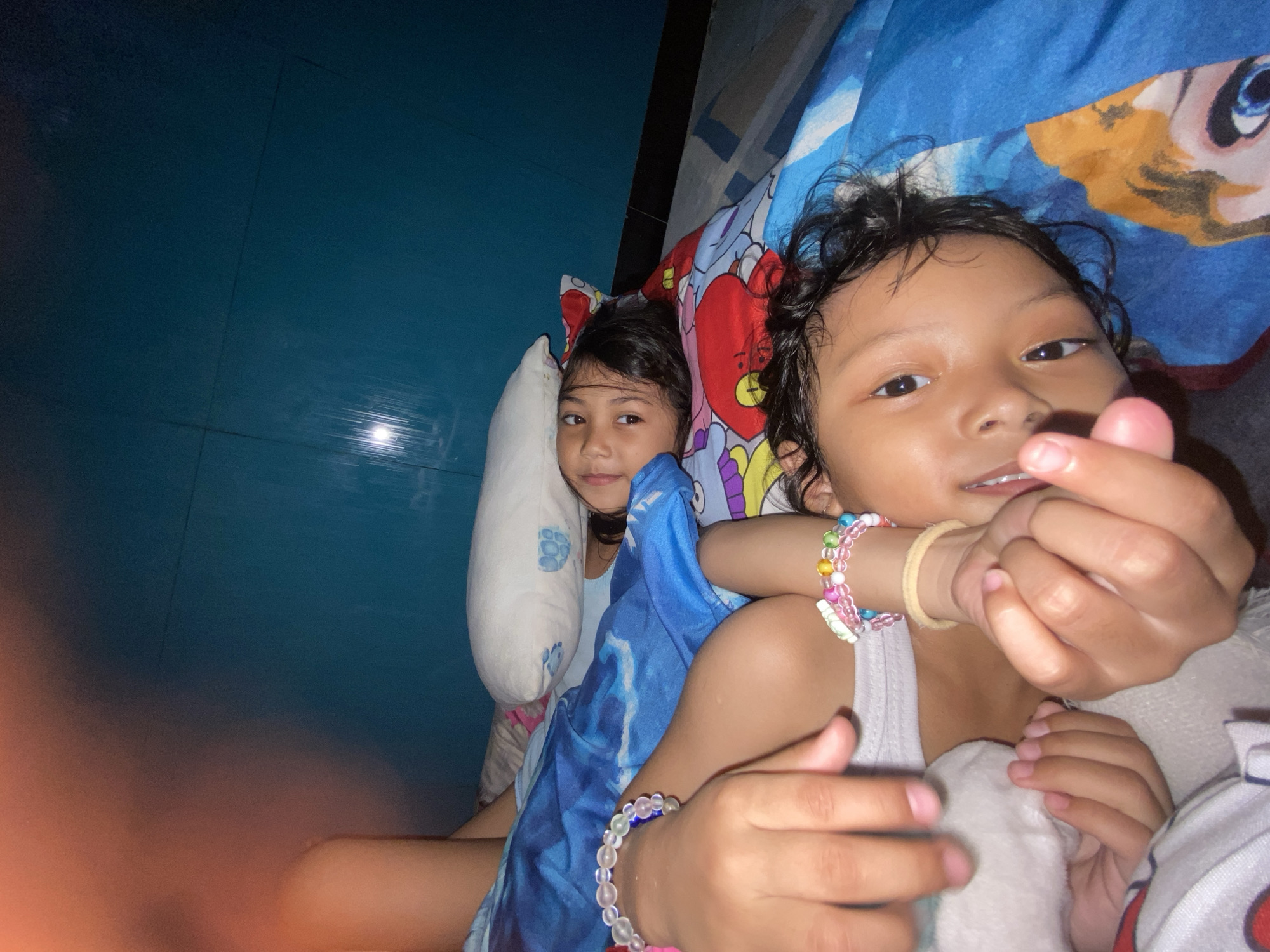 Two young children are lying down in what appears to be a bedroom. One child is behind the other, propped up with a white pillow and blue-patterned fabric. The second child is closer to the camera and is making a heart shape with their fingers. Both children have dark hair and are wearing bracelets. The bedding has a cartoon print. The background is a blue wall. No other information suggests the location is specifically Gelam, Indonesia.