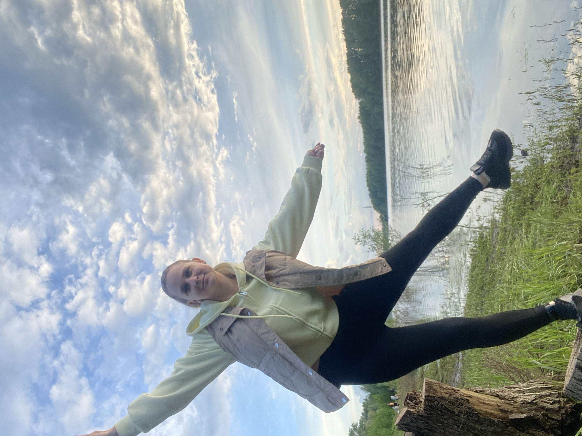 A young woman is posing in a dynamic stance with outstretched arms and legs spread apart, appearing to balance on a log near a body of water. She wears black leggings, black sneakers, a light yellow hoodie, and a beige cropped jacket. The background reveals a lake with a forested shoreline under a sky filled with scattered clouds. The location is the city of Chaikovsky, Russia.