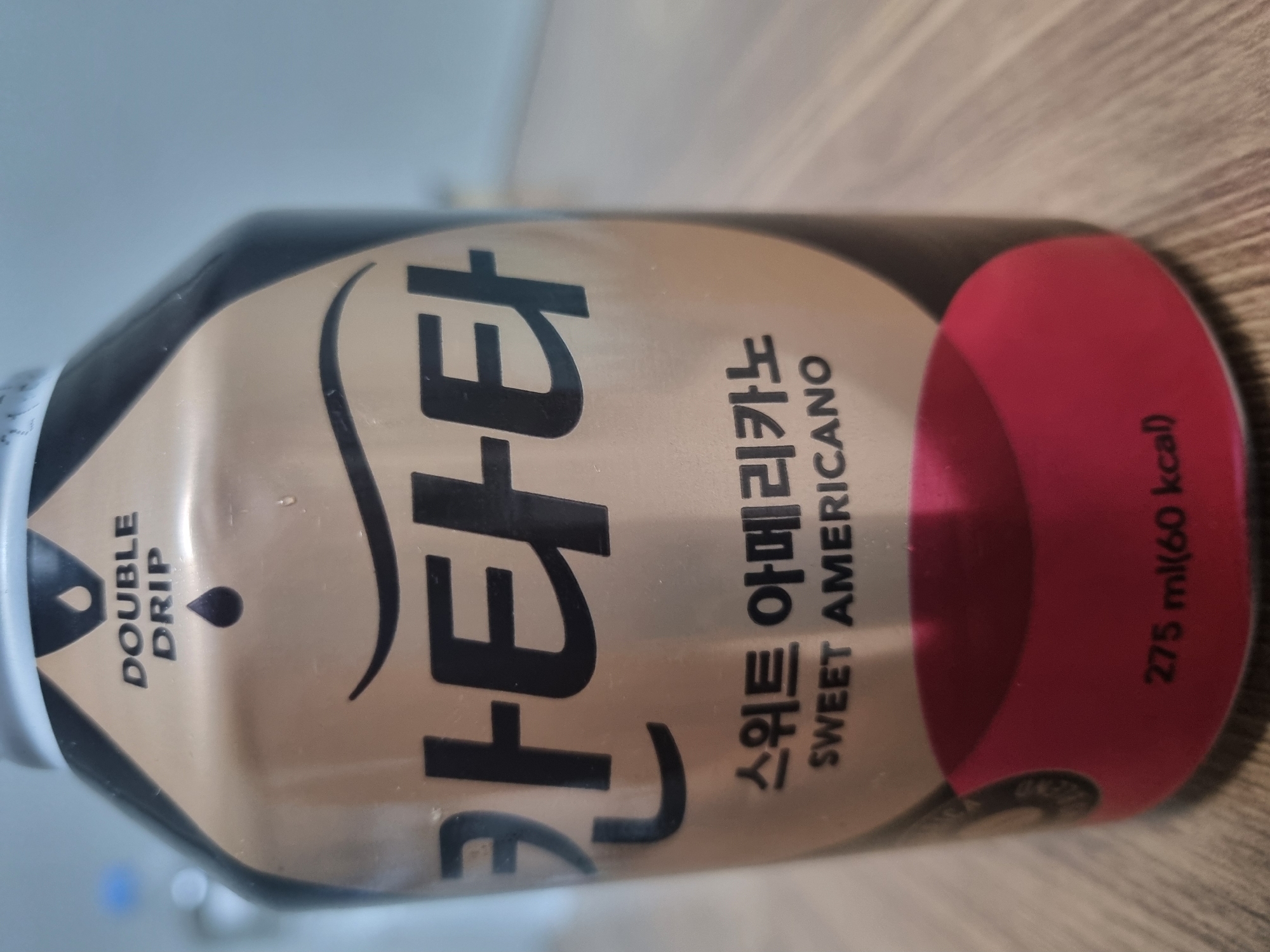 The image shows a close-up of a canned drink on a wooden surface, likely a table. The can is predominantly gold with black text and a red circular design near the bottom. The text on the can is in Korean and English. Prominently displayed in large Korean characters is the brand name "칸타타" (Kantata). Below this, in smaller Korean characters, it reads "스위트 아메리카노" (Sweet Americano), followed by the English translation "SWEET AMERICANO". Near the top of the can, the words "DOUBLE DRIP" are visible, accompanied by an illustration of two water droplets. Towards the bottom of the can, the text "275 ml (60 kcal)" indicates the volume and calorie content. The background is blurred, suggesting a shallow depth of field, and appears to be an indoor setting. There are no people or animals visible in the image. The lighting is even, highlighting the details of the can. This image was captured in Hwaseong-si, South Korea.