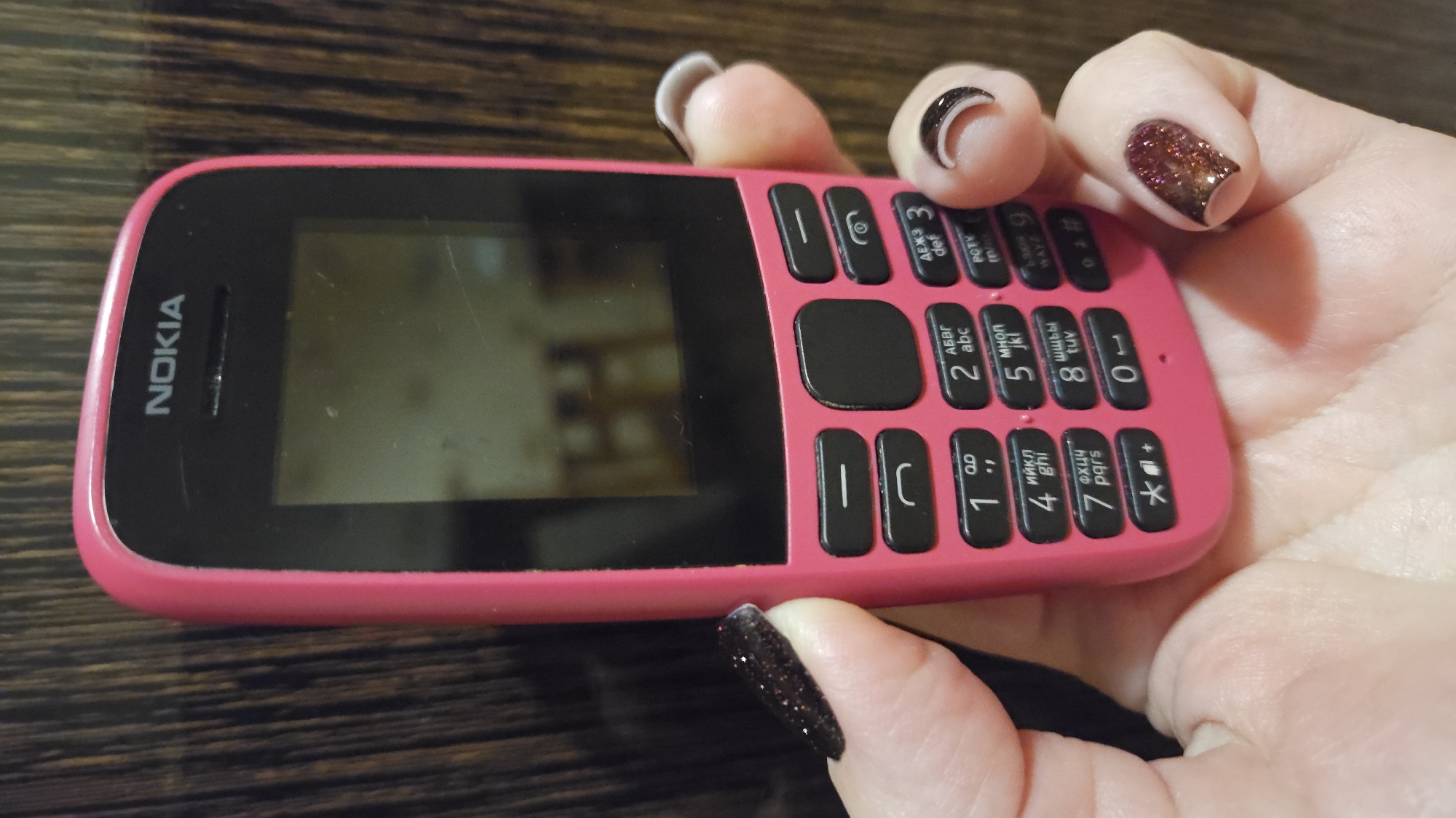 A Nokia feature phone, pink in color, is held in a person's hand. The phone displays Cyrillic characters on the number keys alongside Latin letters. The person's nails are decorated with two different colors: one is a dark glitter with black tips, and the other is a neutral color with dark tips. The background is a dark brown wood surface. The phone has the brand "NOKIA" on the front. The image gives no information regarding Sevastopol or Ukraine.