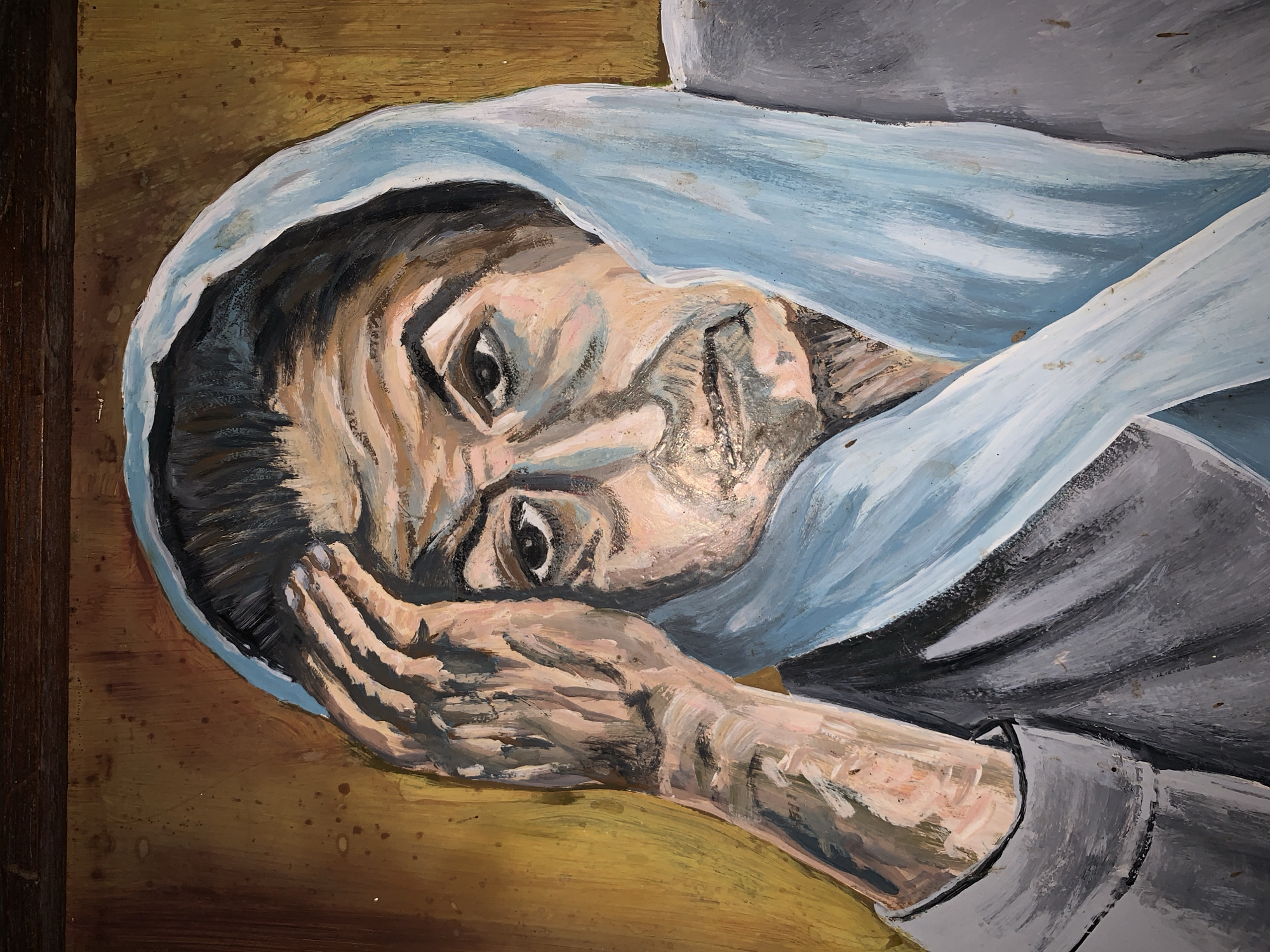 This is a forensic photograph of a painting, likely executed in oil or acrylic, depicting a somber elderly woman in a direct frontal portrait. The woman has a heavily wrinkled face, dark eyes, and a somewhat downturned mouth. She is shown wearing a light blue head covering that drapes over her head and shoulders, and a grey garment. Her right hand is raised to her forehead/temple, with fingers pressed against her skin, suggesting a gesture of contemplation or distress. The painting exhibits thick, visible brushstrokes, especially on the skin tones and the textured background, which features mottled brown and yellow hues. A dark brown, wooden frame element is visible at the top edge of the image. This painting is located in the city of Santa Ana, Venezuela.