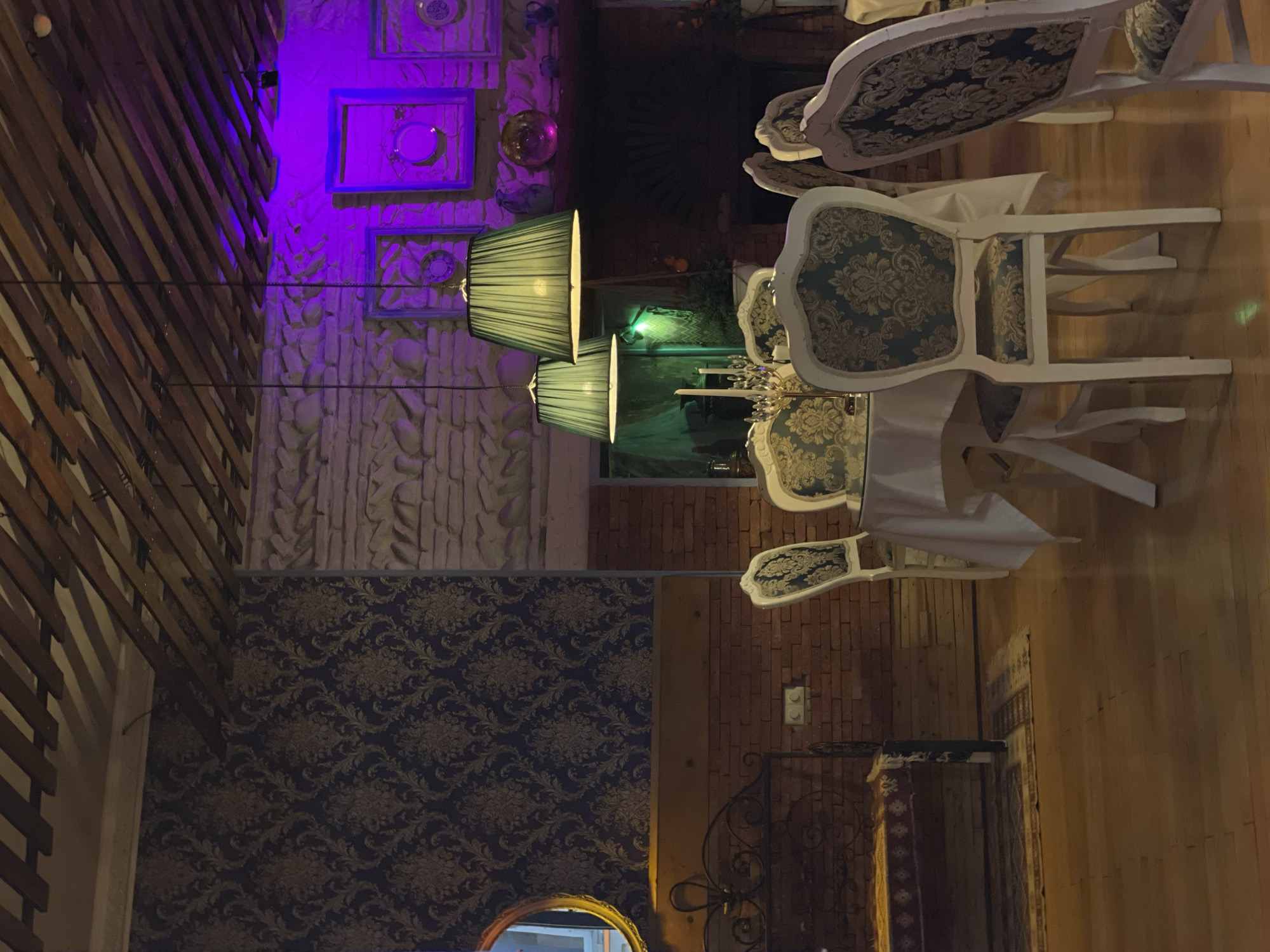 The image shows the interior of a room, likely in a restaurant or similar establishment, in Tbilisi, Georgia. The room is decorated with a mix of styles, including patterned wallpaper, exposed brick, and stone walls illuminated by purple lighting. There is a row of pendant lamps with green shades hanging above a table setting with white chairs, adorned with ornate patterns on their backs. The table is covered with a white tablecloth, and there appear to be decorative objects, possibly candlesticks, on the table. The floor is wooden. A section of a ceiling structure made of wooden slats is visible in the upper left of the frame.