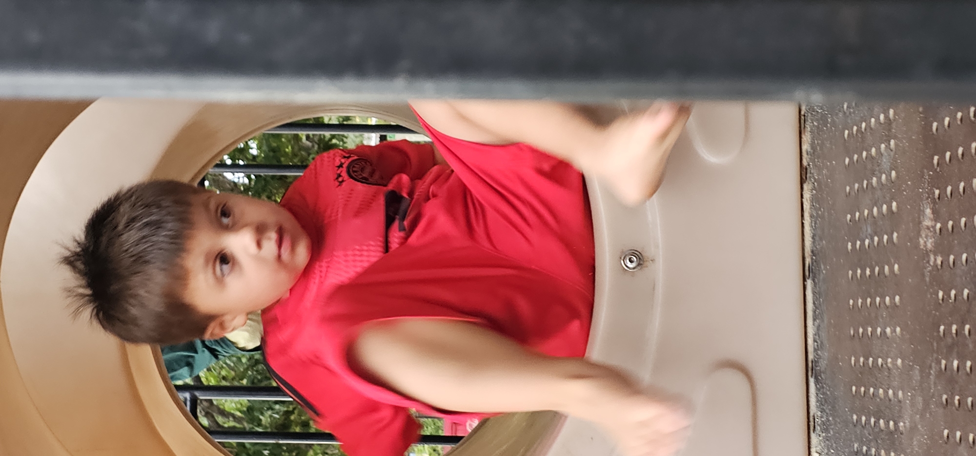A young boy with short, dark hair is captured from an upward perspective, looking directly at the camera while inside a light-beige, cylindrical playground tunnel or slide. He is wearing a bright red short-sleeved shirt with black detailing, including what appears to be a stylized black emblem or crest with three "X"-like symbols arranged vertically on his left chest area, and possibly black stripes on the sleeves.
The boy is positioned dynamically, with his body partially obscured by the tunnel's opening. His legs and feet are in motion, suggesting he is actively playing, possibly crawling or sliding. A metal fastener or bolt is visible on the inner surface of the tunnel below his feet.
The setting is an outdoor playground, evident from the natural light and the background visible through the tunnel's opening. Beyond the beige tunnel, green foliage from trees or bushes and a black metal fence are discernible. To the right of the tunnel's opening, a dark, textured surface, possibly a metal grate or part of another playground element, is visible. Above the boy, a dark, blurry structure, likely part of the playground's upper section, frames the top of the image.
The image appears to be taken during the daytime under bright conditions. The boy's expression is neutral and engaged, showing curiosity as he looks towards the viewer. The original image is oriented sideways, with the boy's head tilted to the right. The provided location context is Denpasar, Indonesia.