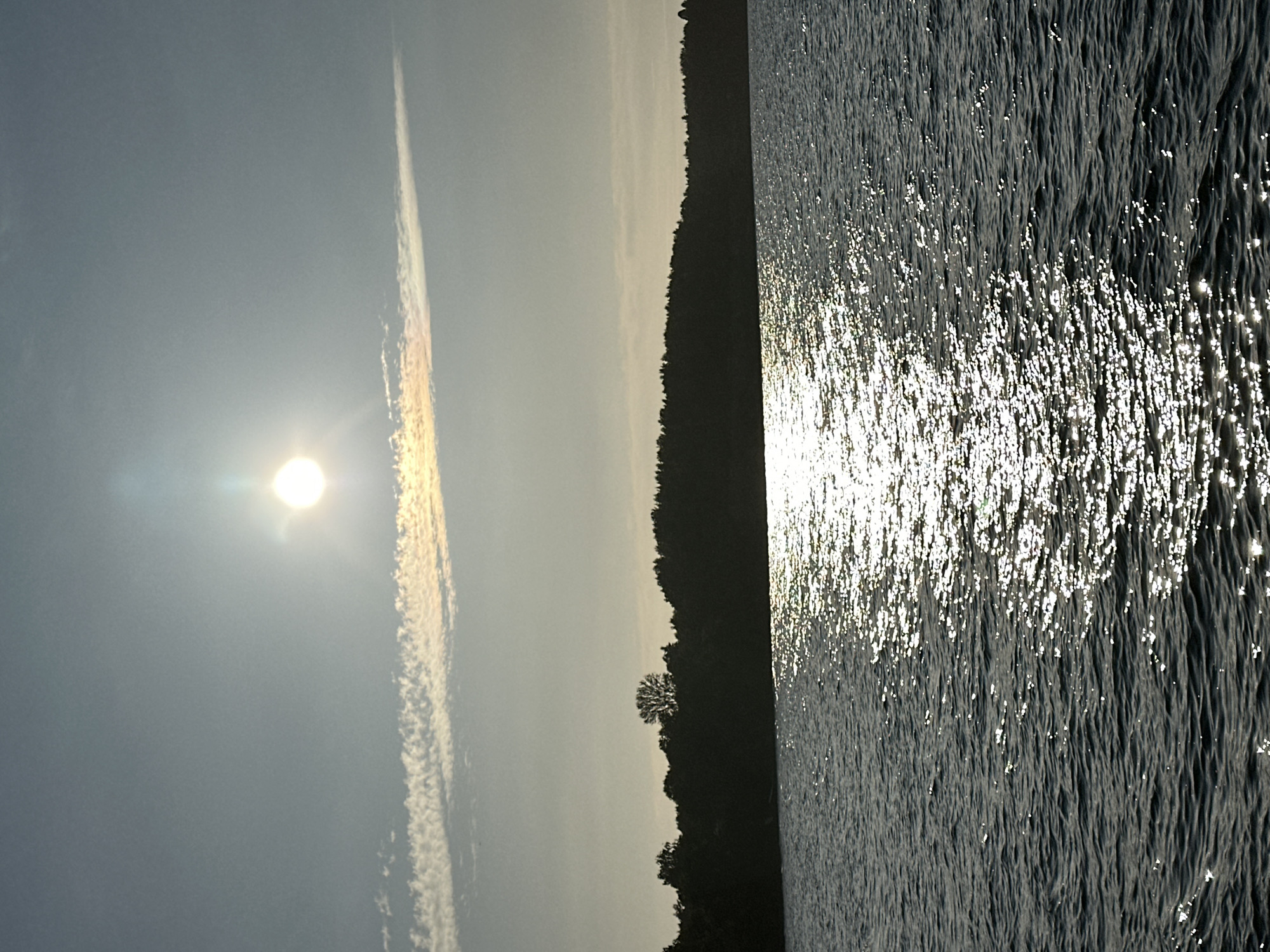 The image shows a wide body of dark water under a hazy sky. A bright sun is visible in the upper portion of the sky, with a thin, streaky cloud below it. The sun's reflection creates a shimmering path across the water's surface. In the distance, a dark, silhouetted treeline runs along the horizon. The water appears calm with small ripples. This scene does not provide any specific details that would allow for identification of a location within Tallinn, Estonia.