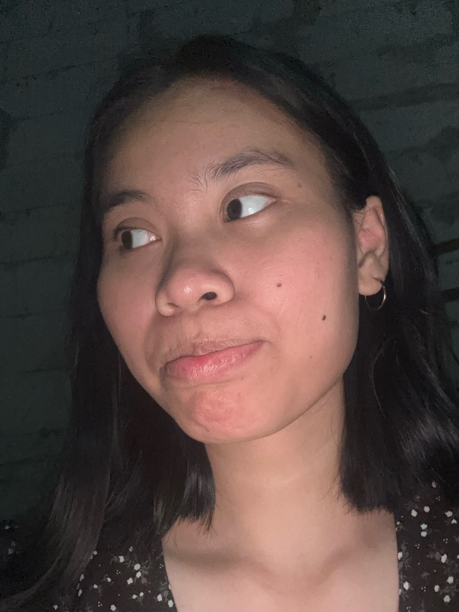 This is a close-up, waist-up, dimly lit, indoor selfie of a young woman. She has dark, shoulder-length hair and appears to be looking off to the left of the frame with a slightly pursed expression. Her skin is fair and smooth, with a few subtle blemishes. She is wearing a dark, patterned top. The background is dark and indistinct, with a textured surface visible on the left, possibly a concrete wall. The overall impression is of a casual, personal photograph taken in a low-light environment. There is no identifiable information regarding the specific location in East Jakarta, Indonesia.