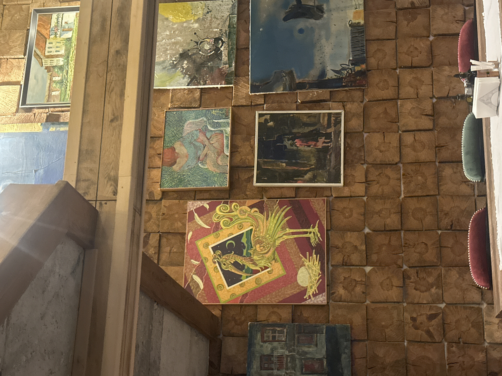 The image shows a wooden wall covered with various framed artworks. Above, a rectangular framed painting depicts an urban landscape with buildings and greenery. Below, a blue-toned painting is partially visible. On the wooden wall, there are four paintings arranged in a square pattern. The top left painting has a yellow and black abstract design. The top right has a blue and gray abstract design. The painting below to the left depicts abstract faces in green tones. The painting to its right is more abstract. Below the square of paintings, there is a painting with an elaborate yellow and green illustration of a mythical creature in a dark sky, surrounded by a red and white border. There is also a small, partially visible painting below it.
The location is Varna, Bulgaria. The wooden walls give the impression of an art gallery or a curated space within a home or public building. There are also green and red pillows visible on the wall to the right of the paintings, suggesting a seating arrangement is nearby. There are other details on the wall to the right as well, including what seems to be a white decorative object on top of the red pillow.
The setting is indoors. The natural lighting indicates that the photo was taken during the day. The environment suggests an artistic or creatively designed space.