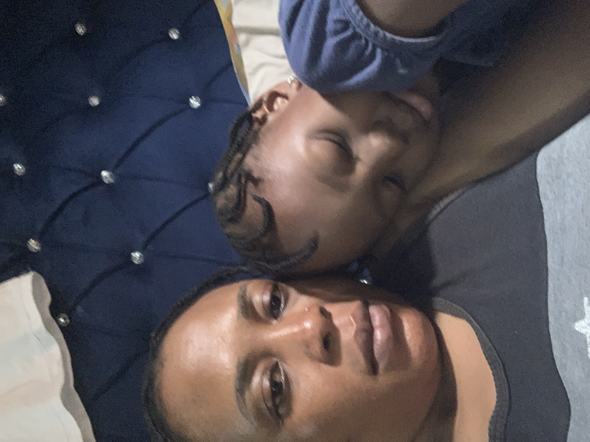 In a bedroom in Sabon Tasha, Nigeria, a Black woman is taking a selfie while a young Black child sleeps on her shoulder, their heads leaning against a dark blue tufted headboard. The woman's eyes are open and looking at the camera, and she has a small silver stud in her nose. She is wearing a dark gray t-shirt. The child is wearing a blue t-shirt and has braided hair. The room is dimly lit.