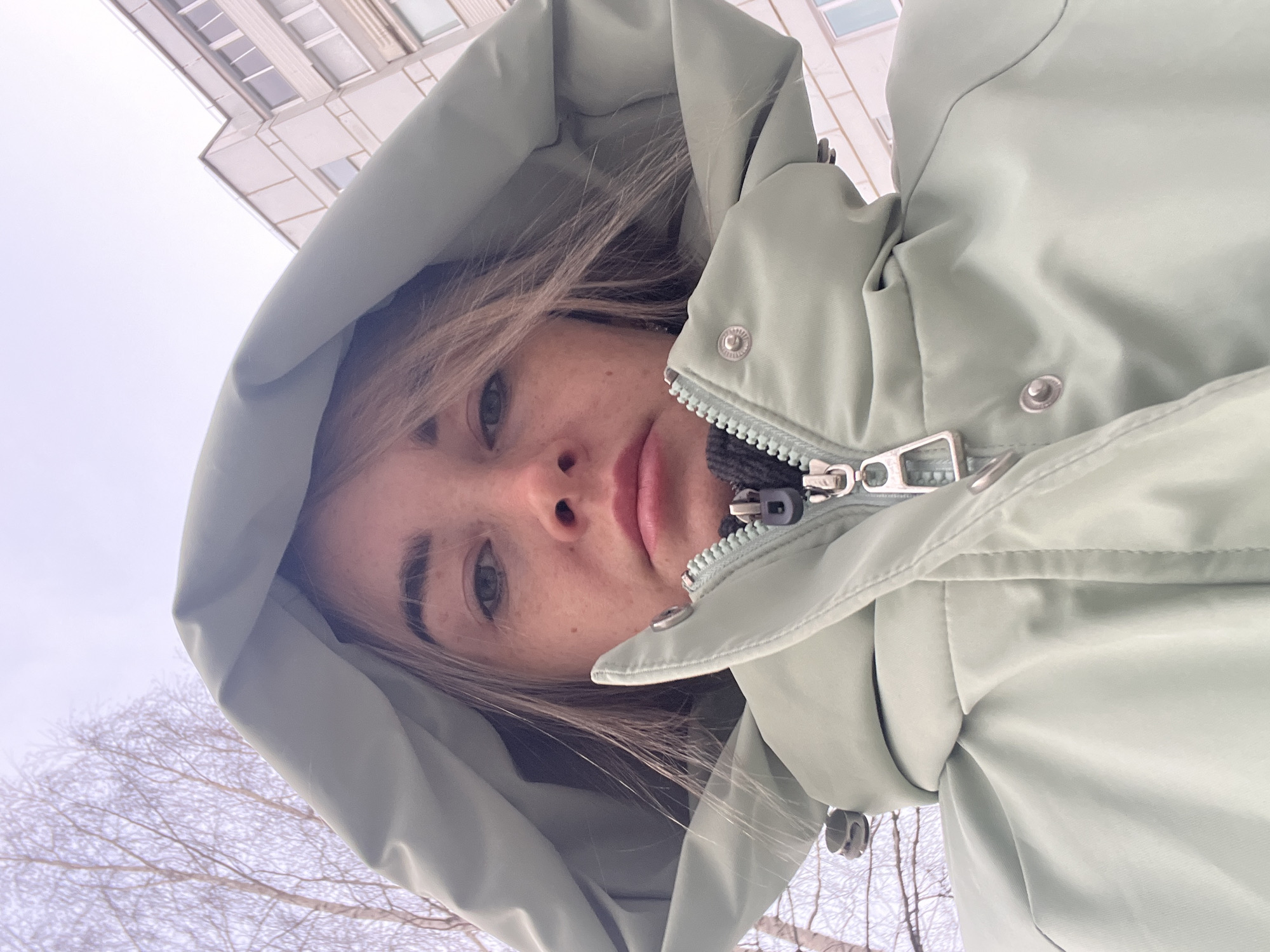 The image presents a low-angle portrait of an adult female in an urban environment within Chaikovsky, Russia. She is directly facing the camera, wearing a light mint-green hooded jacket, partially zipped, revealing a darker garment beneath. Her light-colored hair is visible beneath the hood, and freckles are present on her face. The jacket features a silver-toned zipper pull and metallic snap fasteners. The background on the left side of the frame shows bare deciduous trees under an overcast sky, indicating a colder season. On the right, a segment of a multi-story residential building with light-colored exterior walls and multiple windows is visible, characteristic of urban architecture.