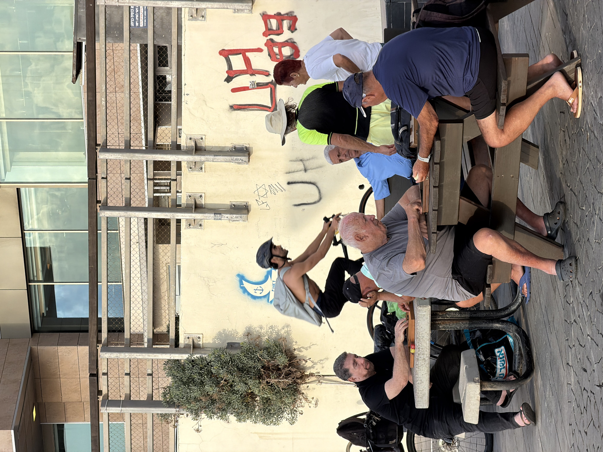 The image shows a group of people gathered around a picnic table in an outdoor setting. The scene appears to be daytime, with bright sunlight casting shadows on the paved ground. The people are a mix of ages and genders, dressed in casual summer clothing. Some are seated at the table, while others are standing or leaning.
One person, wearing a grey t-shirt, is seated at the table and appears to be playing a game with another person. There's a small game board with colorful pieces on the table. Another man, wearing a blue t-shirt and black shorts, is also seated at the table, his legs propped up. A woman with red hair and a white t-shirt is standing behind the table, her head turned away. A man in a black shirt with a baseball cap is visible, leaning forward.
A young man wearing a helmet and a grey backpack is in motion, appearing to be getting on or off a bicycle. He is shirtless and has a determined expression. There is graffiti on the wall behind the group, including red lettering and a blue drawing of a crescent moon with a star. Some of the graffiti appears to be in Hebrew.
The setting seems to be urban, with a building visible in the background and a paved walkway or patio area. The presence of a picnic table suggests a park, public square, or perhaps an outdoor cafe area. The overall atmosphere seems relaxed and social, with the individuals engaged in conversation or activities together.