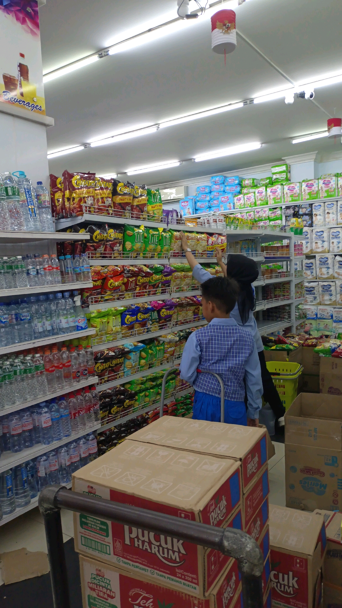The image depicts the interior of a supermarket or minimarket, likely located in Padang Panjang, Indonesia, given the presence of Indonesian text on product packaging. Two individuals, a child wearing a blue and white plaid shirt and blue pants, and a person wearing a black hijab and a blue top, are visible in the foreground, browsing the aisles. They appear to be in the process of shopping or selecting items from the shelves.
The shelves are stocked with a variety of products, including bottled beverages, snacks, and what appear to be diapers or baby products. The lighting is bright, typical of a retail environment, with fluorescent lights overhead. Cardboard boxes are stacked on a trolley and on the floor, some of which bear product branding. The overall scene suggests a typical retail shopping experience.