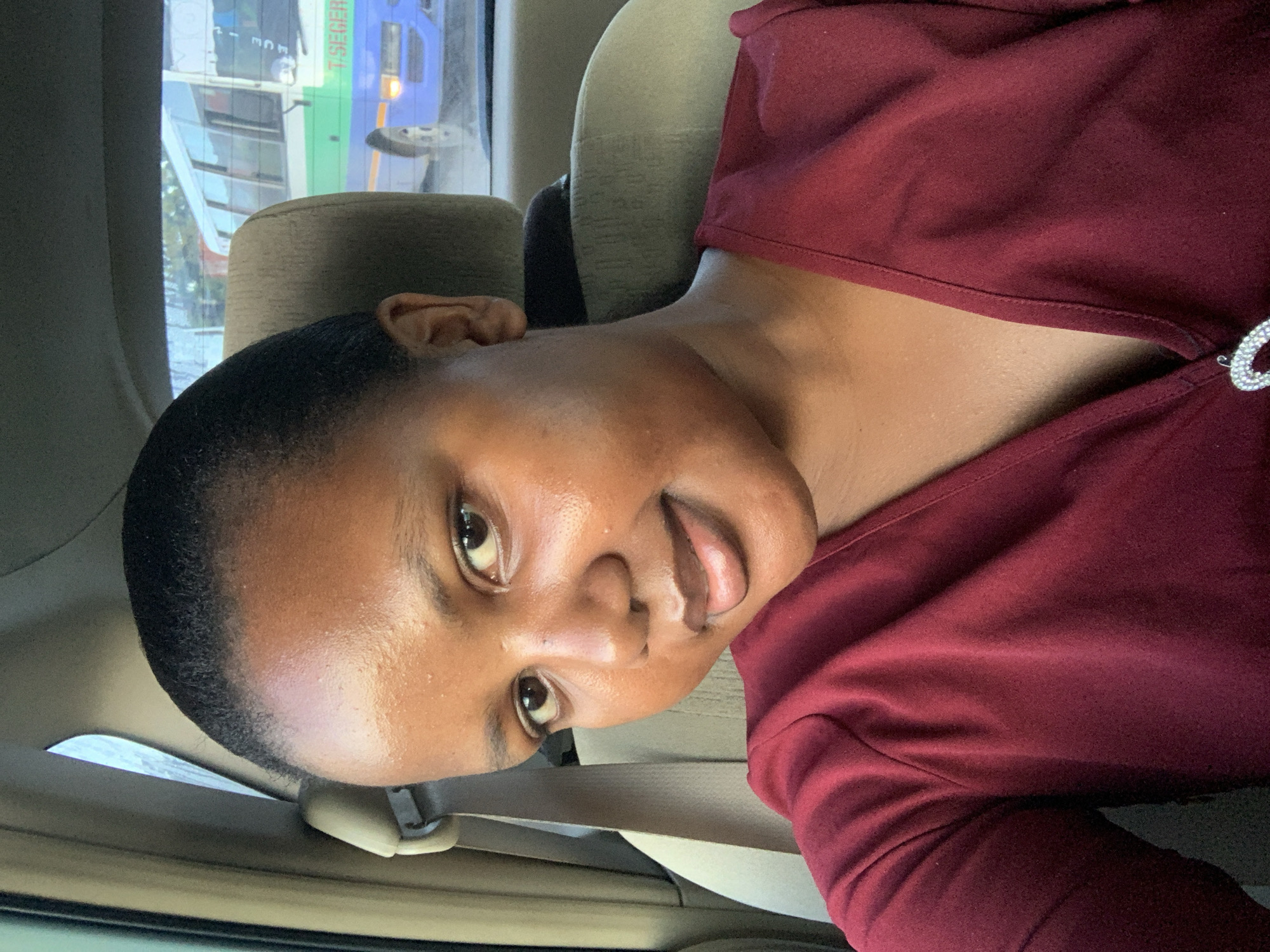 A person with dark skin, short black hair, and wearing a dark red top is sitting inside a car. A public bus is visible through the car window, and the lettering on the bus includes the word "T. SEGERA". Based on the bus information, it's possible that the location is in or near Dar es-Salaam, Tanzania, as T. Segera is likely a local transport company. The person is facing the camera with a slight smile.