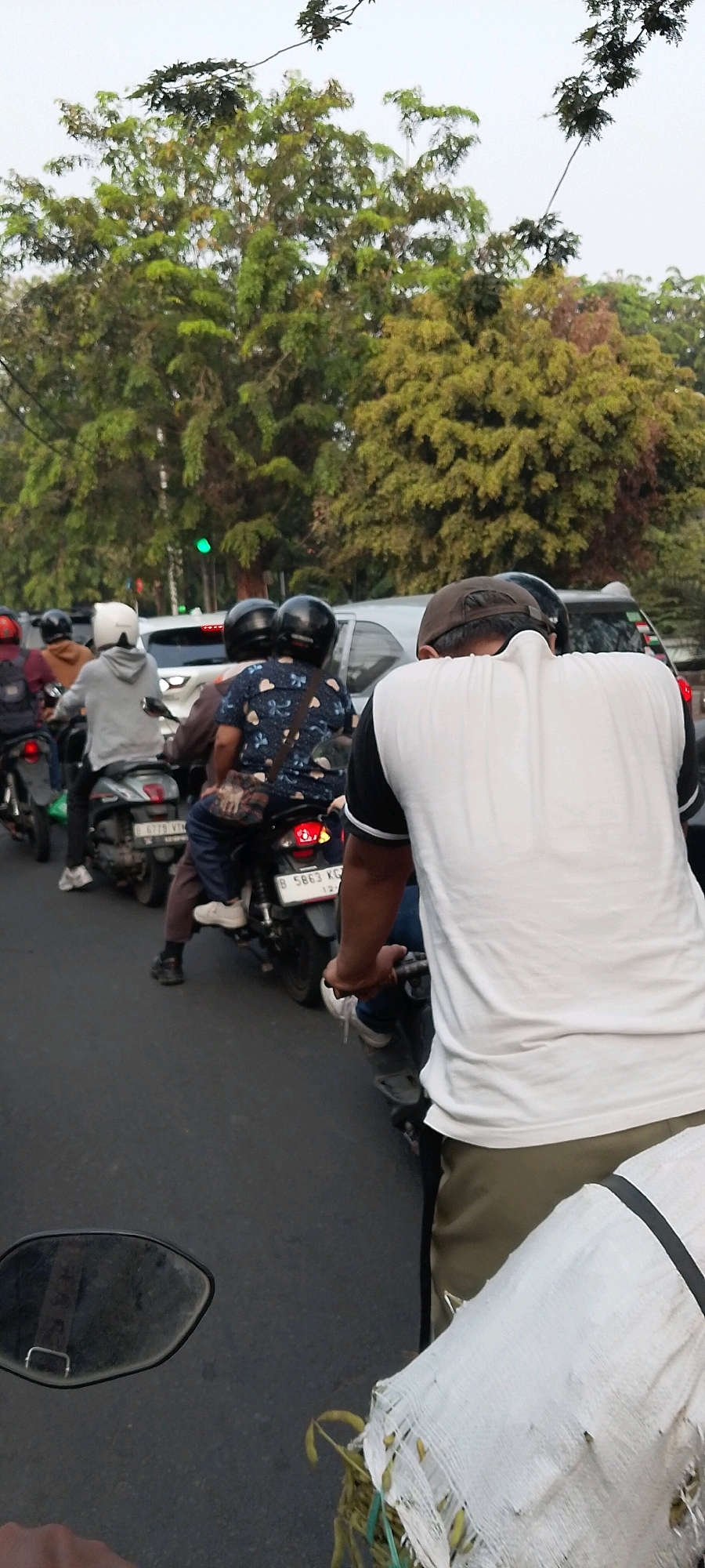 In Bekasi, Indonesia, a traffic jam is depicted on a street lined with trees under an overcast sky. The foreground features the back of a person riding a motorcycle, wearing a white t-shirt with black trim and light brown pants. Their right hand is on the handlebar, and their left arm is visible. Beside them, another motorcycle is seen with its rear light illuminated, carrying a passenger wearing a dark blue shirt with a floral pattern and dark pants. This motorcycle has the license plate "B 5863 KG". To the left of this, another motorcycle is visible with a rider in a grey hoodie and a white helmet. Further to the left, the back of a person in a dark helmet is seen on a motorcycle. In the background, a white car is stopped, and a traffic light displays green. The road surface is asphalt.