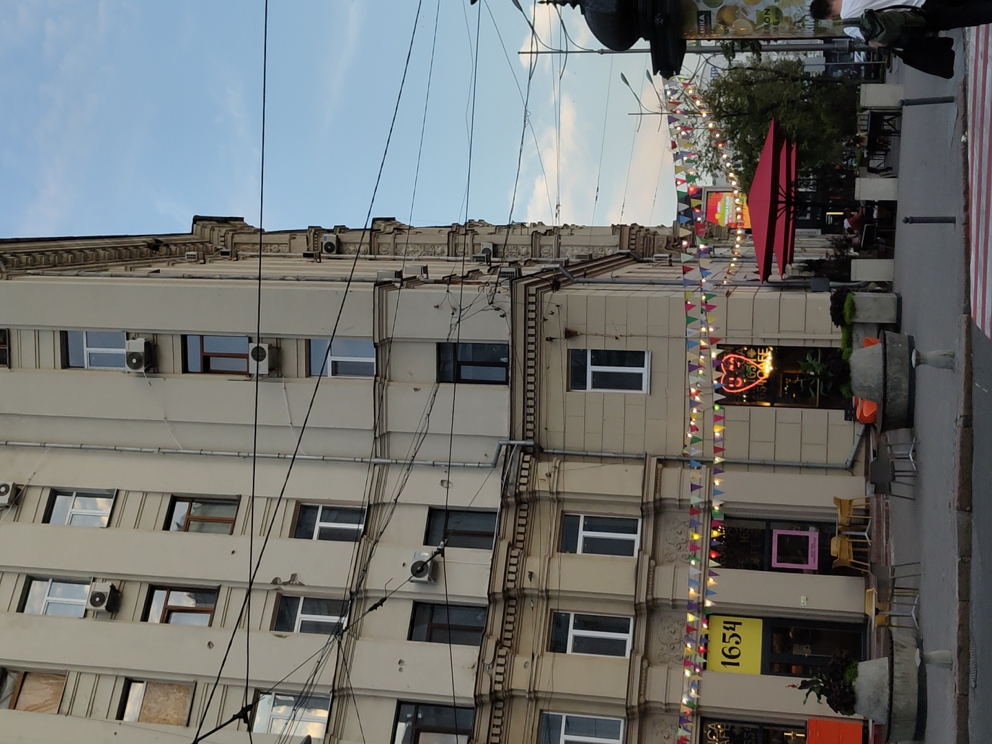 In Kharkiv, Ukraine, a wide shot captures the facade of an older, multi-story building. The building has a light beige exterior and numerous windows, some with air conditioning units. Below, at street level, a cafe named "1654" is visible with a bright yellow sign. Across from the cafe, a red umbrella shades an outdoor seating area. Colorful triangular flags are strung across the street, decorating the scene. The sky above is a pale blue with some clouds. Electrical wires crisscross the view, and a pedestrian crossing is marked in red and white stripes in the foreground. Several people are present, including a person with a backpack walking away from the camera on the right side of the frame, and patrons seated at tables in the cafe's outdoor area. The environment suggests a city street scene in the late afternoon or early evening.