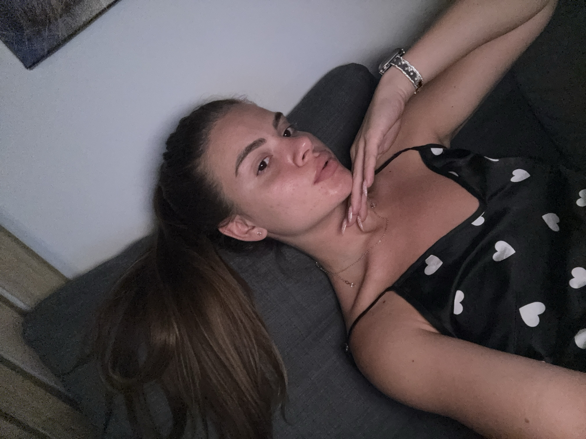 A young woman with light brown hair tied in a ponytail is lying on a dark gray sofa, propped up against a light gray wall. She is wearing a black slip dress with a white heart pattern. Her right arm is bent and resting on her chest, with her hand touching her face. She has on a watch with a diamond-studded band on her left wrist and a delicate necklace around her neck. The woman is looking to her left, with her lips slightly parted. In the background, a framed picture is visible on the wall. The image appears to be taken indoors, possibly in a residential setting. The location is not specified within the city of Plovdiv, Bulgaria.