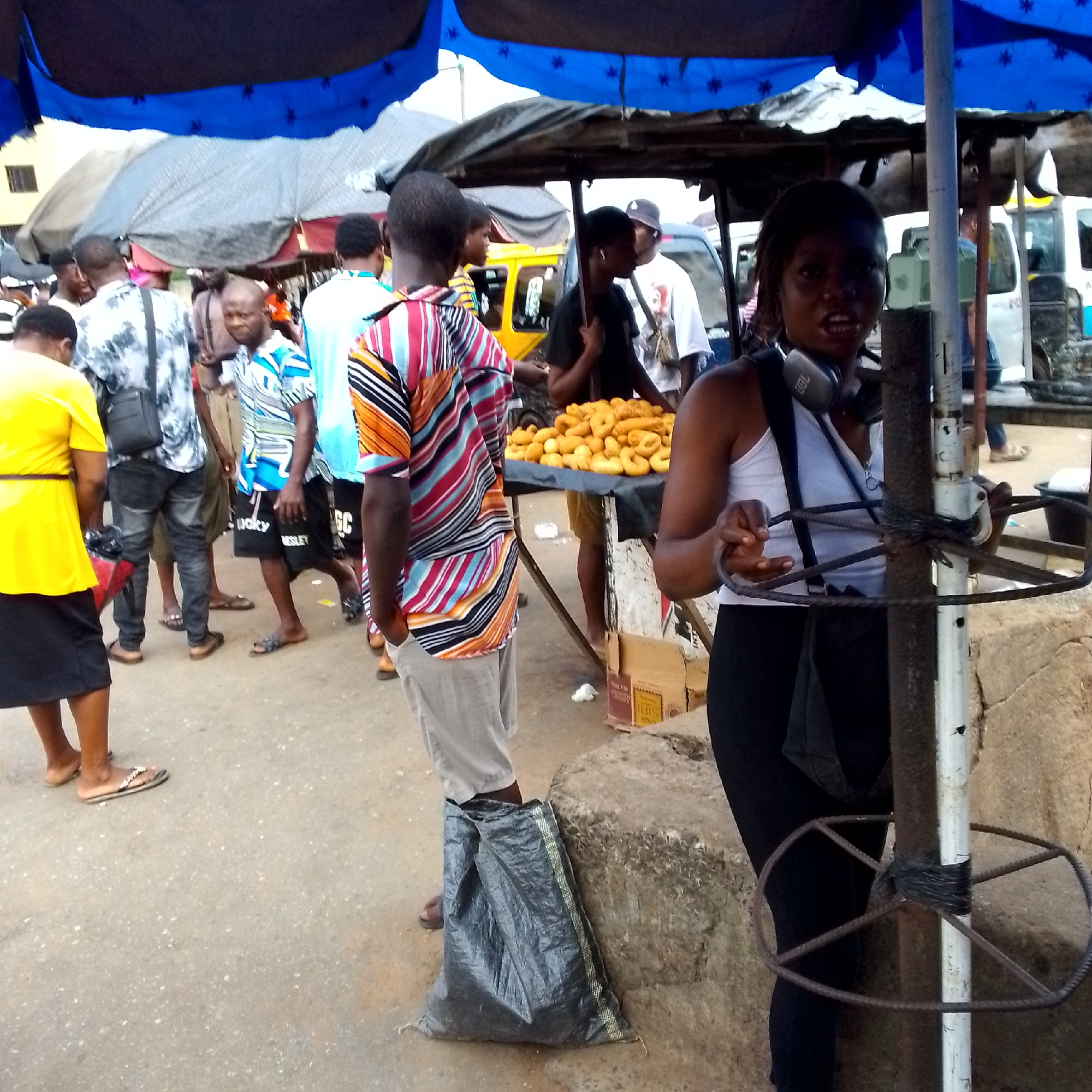 The image depicts a bustling outdoor market scene in Nkpor, Nigeria, likely on a paved road surface. A vibrant blue and black awning stretches overhead, providing shade for stalls and shoppers. In the foreground, a young woman with headphones around her neck, wearing a white tank top and black pants, stands near a metal pole wrapped with black tape and adorned with a circular metal structure. She is looking towards the camera with an open mouth. To her right, a table displays a mound of golden-brown fried pastries. Further into the scene, several other individuals are visible, including a man in a colorful striped shirt, a person in a yellow top, and others dressed in various casual attire. The background reveals more market stalls, a yellow vehicle resembling a tricycle taxi, and another larger white vehicle, possibly a bus. The atmosphere appears lively and commercial, with people engaged in activities typical of a Nigerian market.