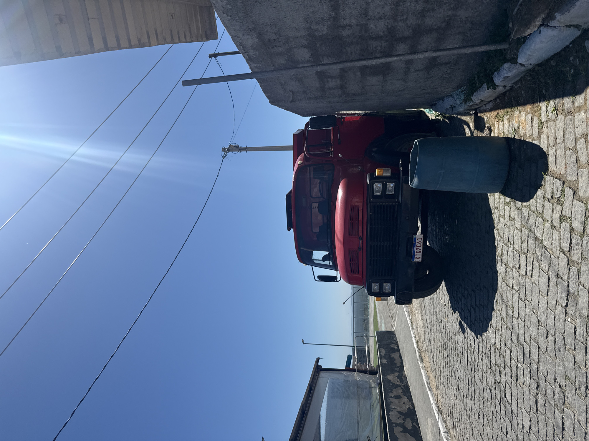 A red truck is parked on a cobblestone street next to a tall, rough-textured concrete wall. The truck, a model likely from the mid-20th century, has its front facing the viewer and the driver's side door slightly ajar. A large, teal-colored plastic barrel stands on the cobblestones to the left of the truck's front tire. Overhead, several electrical wires crisscross against a bright, clear blue sky, with the sun creating lens flare in the upper right portion of the frame. To the left of the truck, a modern-looking building with a clear plastic roof and a white base is visible. The street slopes slightly downwards, and in the distance, a hazy landscape with some vegetation can be seen under the expansive sky. The overall scene appears to be a quiet, possibly residential or commercial street in Arraial Do Cabo, Brazil, given the style of the truck and the general outdoor setting.