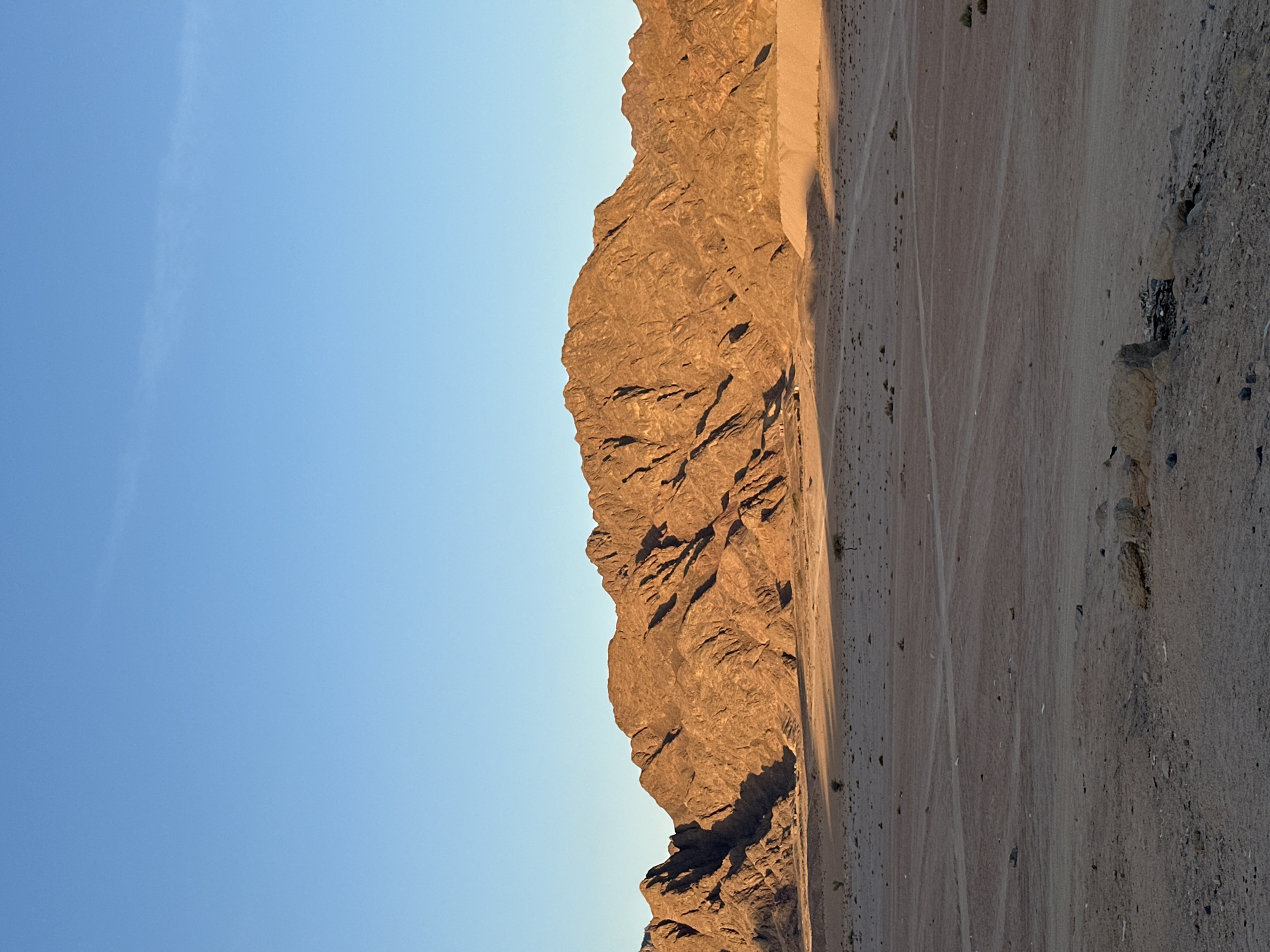 The image shows a mountain range adjacent to a sandy plain under a clear blue sky. The mountain is illuminated by the sun, highlighting its craggy texture. The sandy plain shows tire tracks and scattered rocks. The scene is located in Sharm El Sheikh, Egypt, given the characteristic landscape.