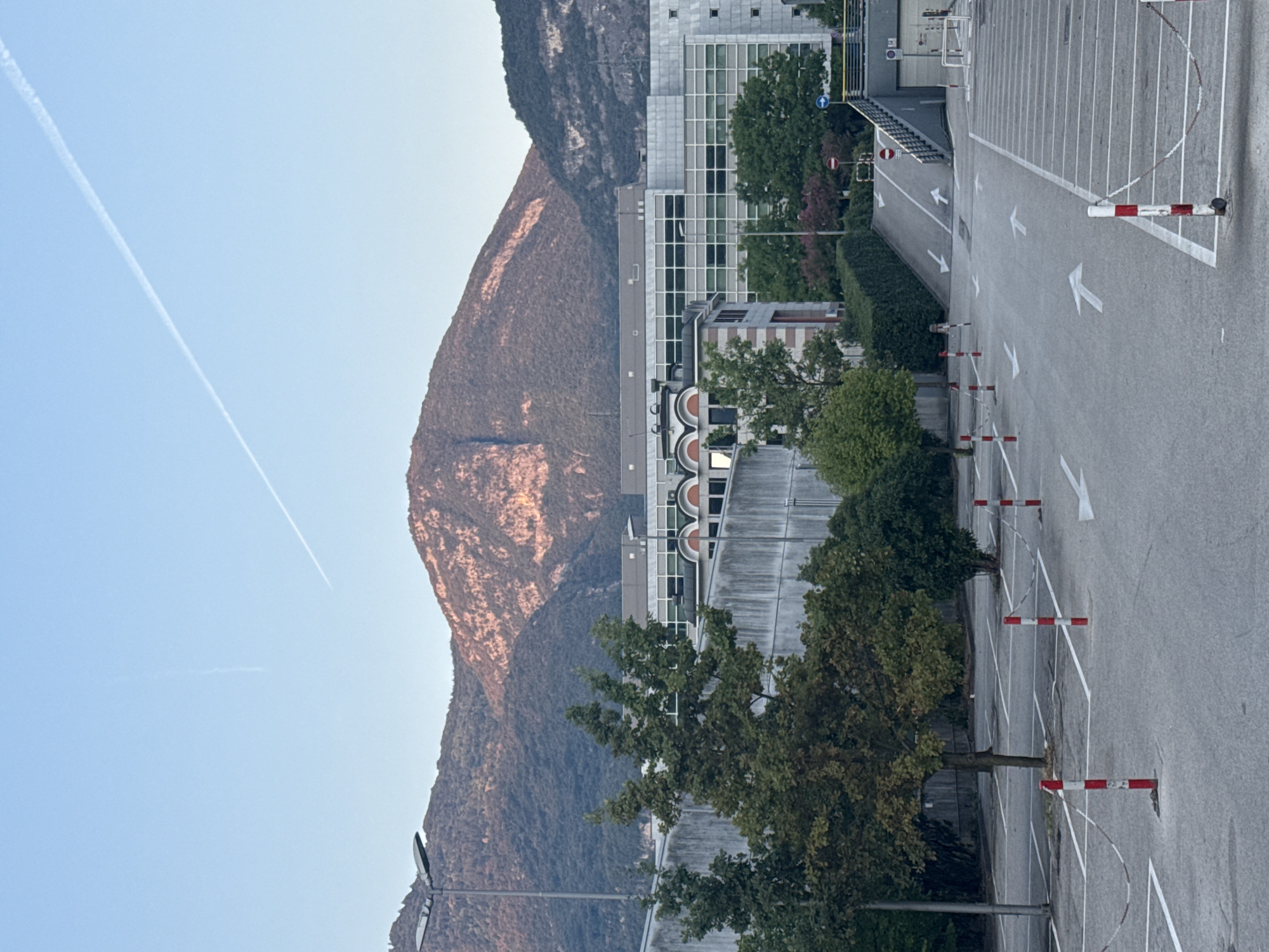 The image depicts a view of a parking lot in front of a modern building, with mountains in the background. The sky is clear and blue, with two faint contrails from airplanes visible. The mountains are covered in trees and show hints of sunlight on their peaks. The building appears to be an office or commercial complex, with large windows. In the foreground, the asphalt parking lot is marked with white lines indicating parking spaces and arrows showing the direction of traffic. Several red and white striped bollards are placed throughout the parking lot, some connected by chains. Trees and some bushes are also present in the scene. The image appears to have been taken in the late afternoon or early morning, given the lighting. This scene is consistent with an industrial or business park area on the outskirts of Trento, Italy, possibly near the north side of the city given the mountainous backdrop. There are no people or vehicles visible in the image.