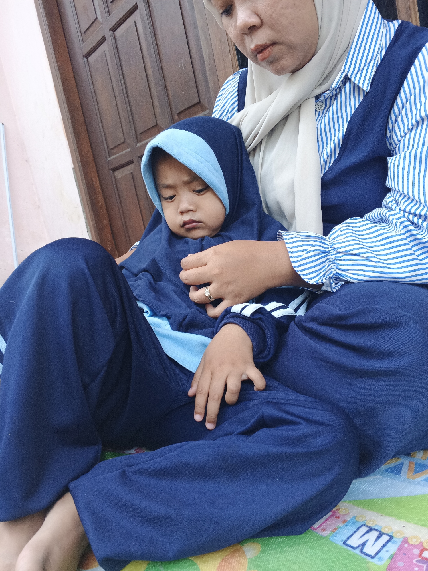 A person wearing a hijab and a blue and white striped shirt sits on the ground, holding a child wearing a blue hooded outfit. The child is looking down with a serious expression. The background shows a wooden door and a pink wall, suggesting an outdoor setting. The ground beneath them is covered by a mat with colorful letter shapes. The image appears to be taken in Warungkiara, Indonesia, as indicated by the provided context.