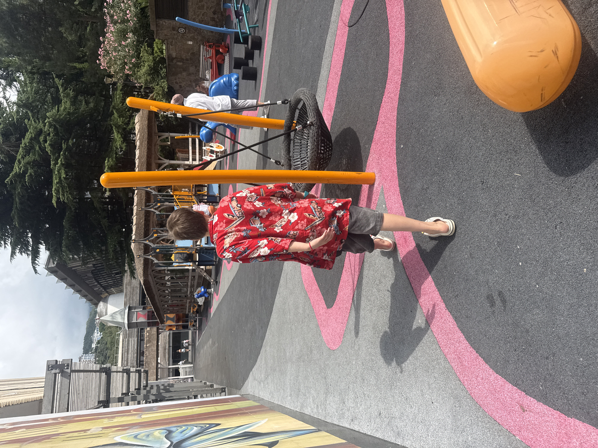 The image shows an outdoor playground area with a young person walking away from the camera. The person is wearing a red Hawaiian shirt and gray shorts. The playground surface is black with pink wavy lines. In the background, there are various playground structures, including swings and climbing equipment, along with some trees and buildings. The architectural style of the background buildings suggests a location like Yalta, Ukraine.