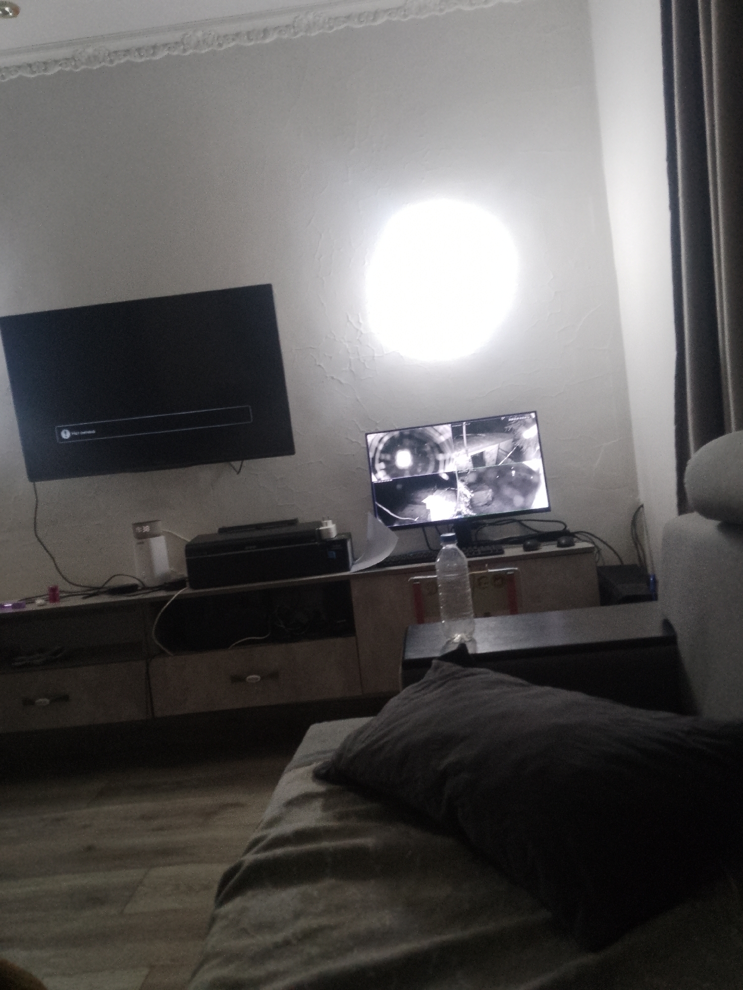 The image depicts an indoor scene, likely a living room or entertainment area, possibly in a residential setting. There are two televisions visible. One is a large, flat-screen TV displaying a loading or menu screen. Below this TV is a printer and other electronic devices. A second, smaller TV is situated to the right, displaying multiple camera feeds, suggesting a security system. A water bottle is placed on a surface in front of this second TV. The room has textured walls with a bright, circular light source on the wall behind the TVs, possibly a lamp or a reflection. In the foreground, a dark-colored pillow rests on a couch or bed. The overall lighting is dim, with the exception of the bright light source on the wall and the screens of the televisions. No individuals are visible in the image, nor is there any direct evidence to suggest the location is in the city of Келдемұрат, Kazakhstan. The scene appears to be a still-life arrangement of electronic equipment and furniture.