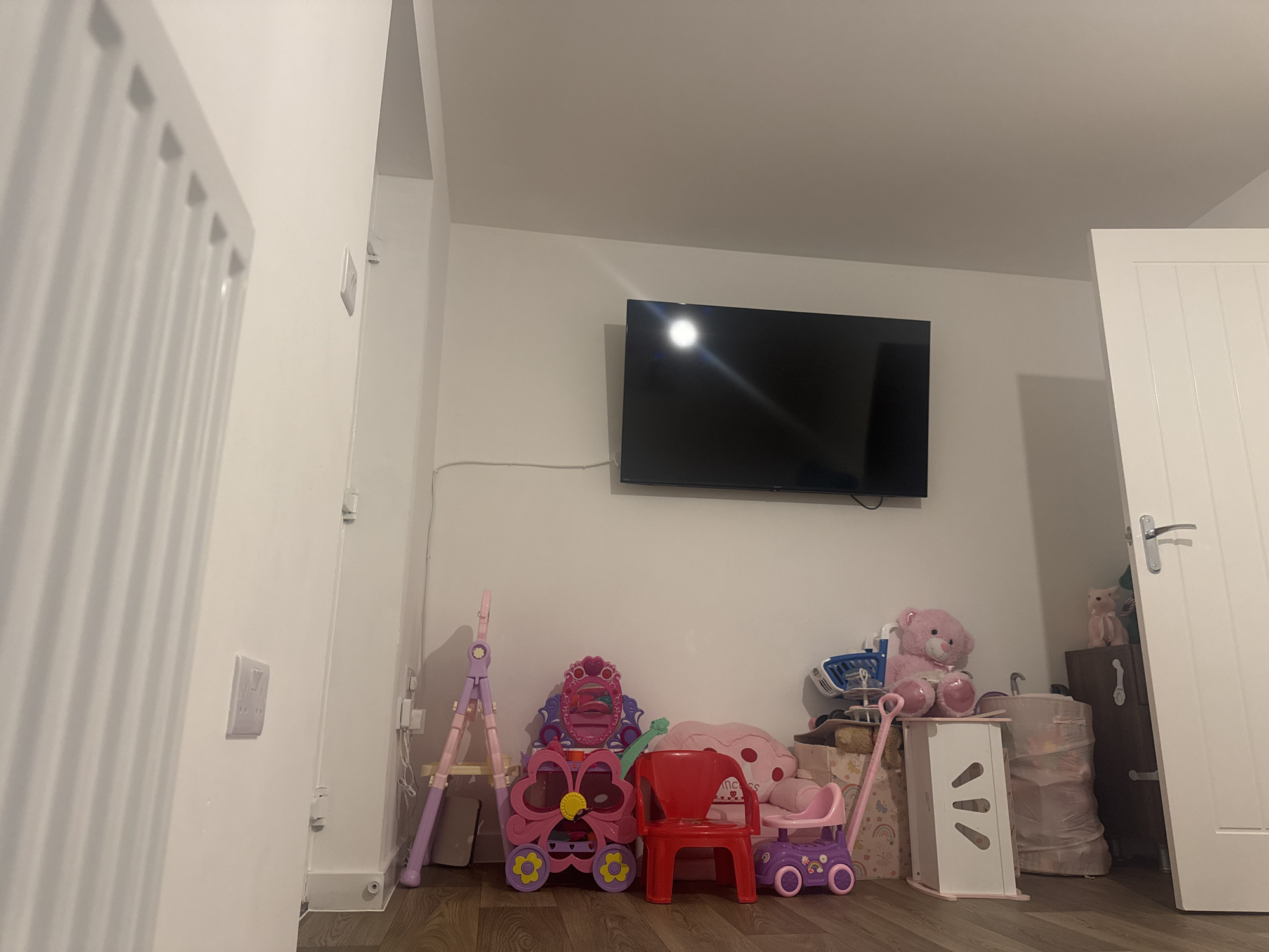A photograph taken indoors depicts a room with various objects, most notably children's toys. The room features white walls and a dark wood floor. On the wall hangs a flat-screen television. In front of the wall, a collection of toys is arranged including a pink play vanity, a red plastic chair, a purple walker toy, and a pink teddy bear. A white storage unit stands to the right of the toys. In the corner, near a white door, there's a cabinet with a couple of stuffed animals atop it. The setting appears to be a playroom or child's bedroom. The location is the Vale of White Horse, United Kingdom. No people are visible in the photograph.