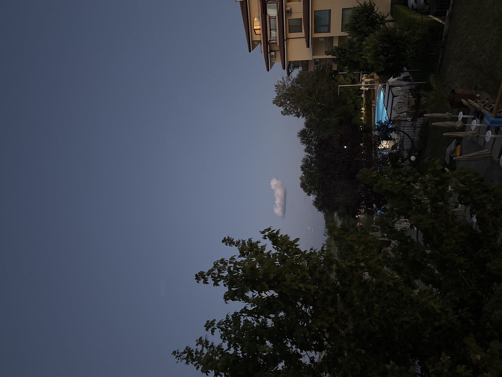 This image depicts a twilight scene in Nesebar, Bulgaria, likely in a resort area. In the foreground, dark green trees with dense foliage dominate the left side of the frame. Beyond the trees, a well-lit swimming pool with bright blue water is visible, surrounded by lounge chairs and umbrellas. To the right, a multi-story building with a yellowish facade and reddish-brown roof is prominent, featuring balconies and illuminated windows. The sky is a deep blue, indicating dusk or early night, with a single small cloud and a distant pinpoint of light, possibly a star or airplane. The overall atmosphere is serene, transitioning from day to night.