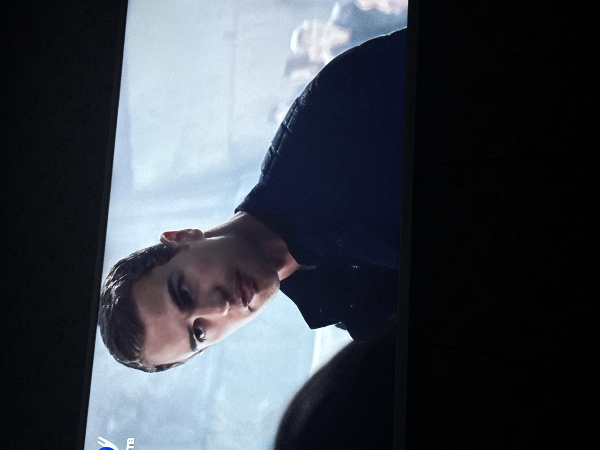 A man is on a television screen. He has short brown hair and is wearing a dark jacket. The background is a light gray. The bottom left corner of the screen has a logo with "ТВ" beside it. The surrounding environment is dark. I cannot confirm the city of Mytishchi, Russia based on the image.