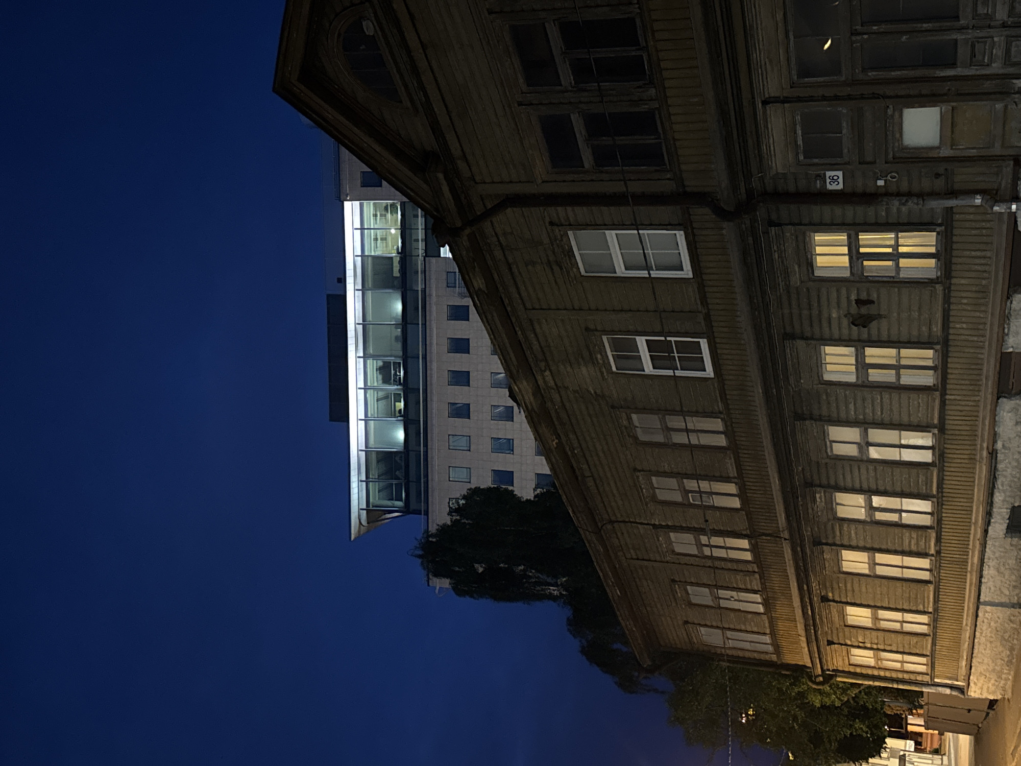 This image depicts a street scene in Tallinn, Estonia, at dusk or night. In the foreground, a dark brown, weathered wooden building with multiple windows is visible. The windows on the lower level are illuminated from within, casting a warm yellow glow. The upper level of the wooden building also has windows, some of which are dark, while others show faint interior light. A downspout runs vertically along the side of the wooden building. A small sign with the number "36" is affixed to the building above one of the ground-level doorways.
Behind the wooden building, a taller, modern building with a glass facade is visible. This structure is brightly lit from within, with rows of windows showcasing the illuminated interior. The sky above is a deep, clear blue, indicating either late evening or early morning. Trees with dark foliage are present to the left of the wooden building, partially obscuring the view of the modern structure. The overall impression is a juxtaposition of old and new architecture in an urban setting at night.
