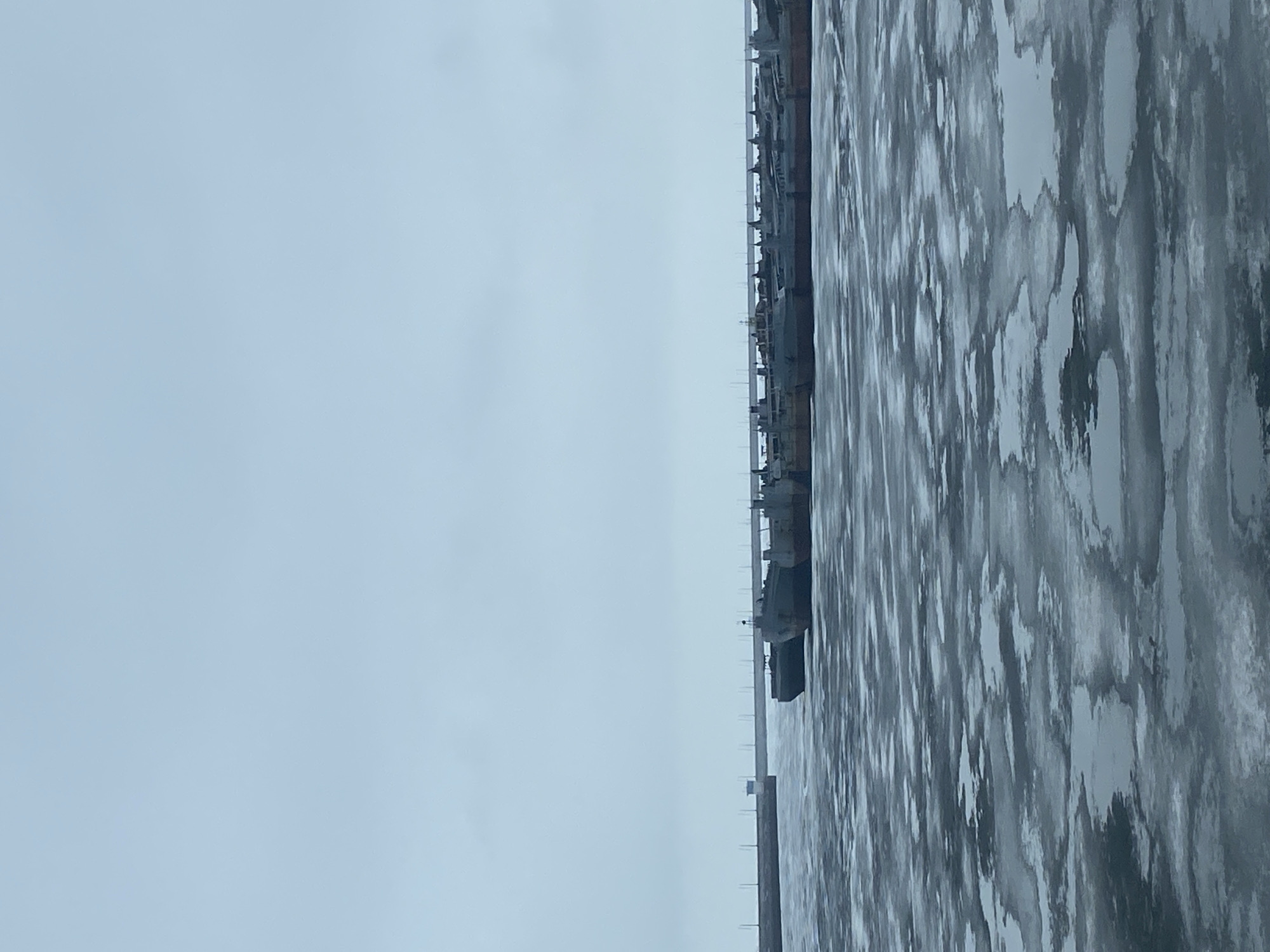 The image shows a pier with ships docked along it, adjacent to a body of water that appears to be partially frozen with ice floes. The sky is overcast and grey. This scene is located in Chaikovsky, Russia.