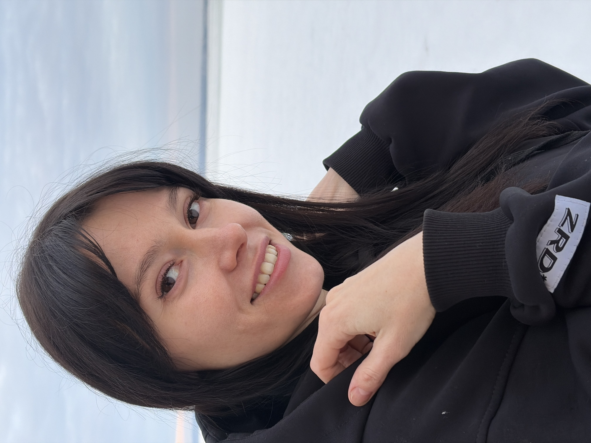 A forensic description of the scene in Noviy, Russia, depicts a female subject outdoors in a winter environment. The subject is a young to middle-aged adult woman with long, dark hair and fair skin. She is positioned slightly off-center to the left, facing forward with a gentle smile, revealing her upper teeth. Her dark eyes are directed towards the viewer. She wears a black hooded sweatshirt with ribbed cuffs. Her left hand is raised to her neck area, grasping the fabric of the hoodie collar or hood. A white fabric tag with black text, partially displaying "ZRD" and an asterisk, is visible on the ribbed cuff of her right sleeve. The background consists of a vast, flat, snow-covered or ice-covered expanse, suggesting a frozen landscape or body of water. A faint horizon line separates the white expanse from an overcast, light blue-grey sky. The lighting is soft and diffused, indicative of natural daylight.