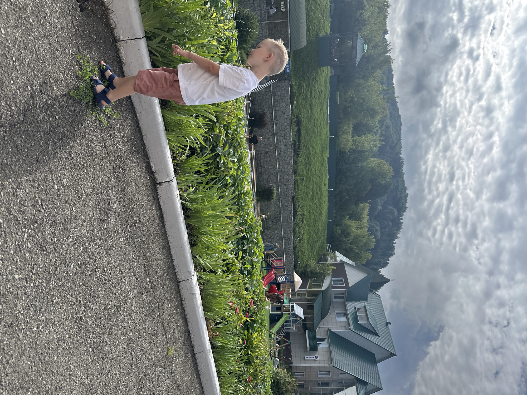 The image depicts a young blond boy in a white t-shirt and pink shorts, wearing dark blue sandals, standing on an asphalt surface near a curb and a flowerbed. He is looking down towards the plants. In the background, a large building with a green roof is visible, along with a playground with slides and climbing structures. Further in the distance, a grassy hill slopes upwards, covered with trees. The sky is cloudy. The scene appears to be an outdoor recreational area, possibly a park or resort, in Даниловка (Алтайский), Russia. The overall atmosphere is casual and suggests a daytime setting.