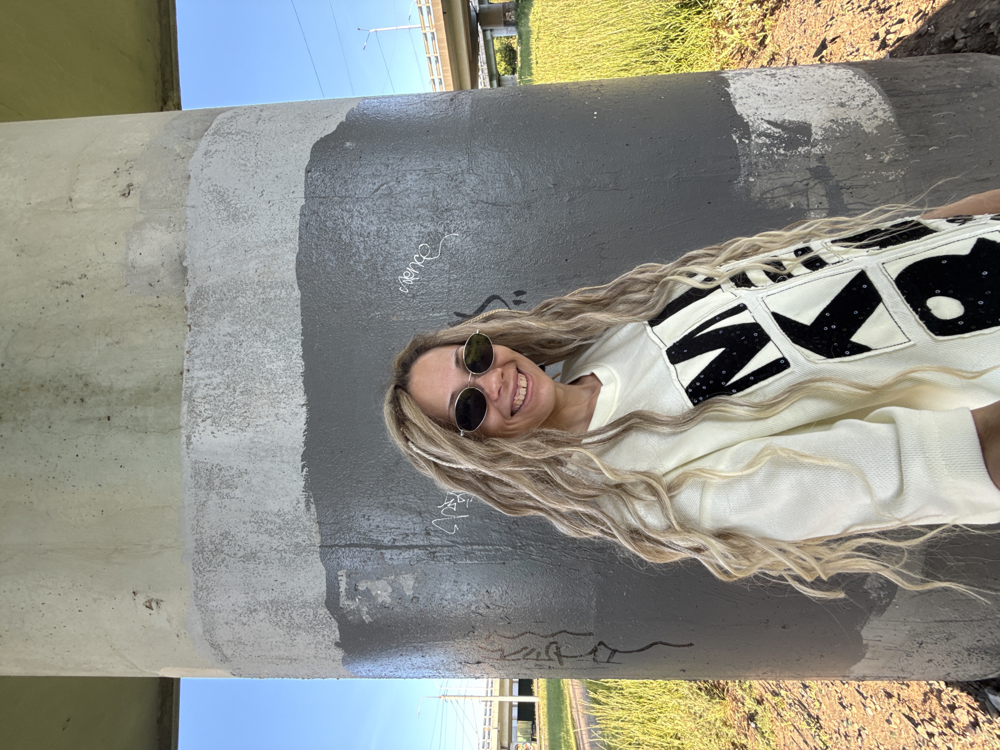 A female subject is depicted standing outdoors on a sunny day in Заволжье, Russia, positioned against a large concrete pillar. The subject has long, wavy blonde hair with integrated dreadlock-style braids, and is wearing dark, oval-framed sunglasses. She is dressed in a light-colored (off-white or cream) long-sleeved sweater adorned with large black letter patches, including partially visible letters "M", "K", and "R". Her facial expression conveys a smile, with her teeth visible.
The concrete pillar occupies the central and right portions of the frame. Its surface exhibits multiple layers of finish: an upper section of exposed light grey concrete, followed by a band of lighter grey paint, and a dominant lower section coated in dark grey, glossy paint. White paint patches are present over the dark grey section, along with various graffiti markings, including a legible "spence" in white script.
To the left of the pillar, a grassy area with tall, yellow-green vegetation is visible, extending towards a road bridge structure with railings and utility poles in the mid-ground. A clear blue sky is apparent above the bridge. To the right of the pillar, similar grassy vegetation extends into the background, with another bridge structure visible further in the distance. The scene is brightly illuminated by natural sunlight.