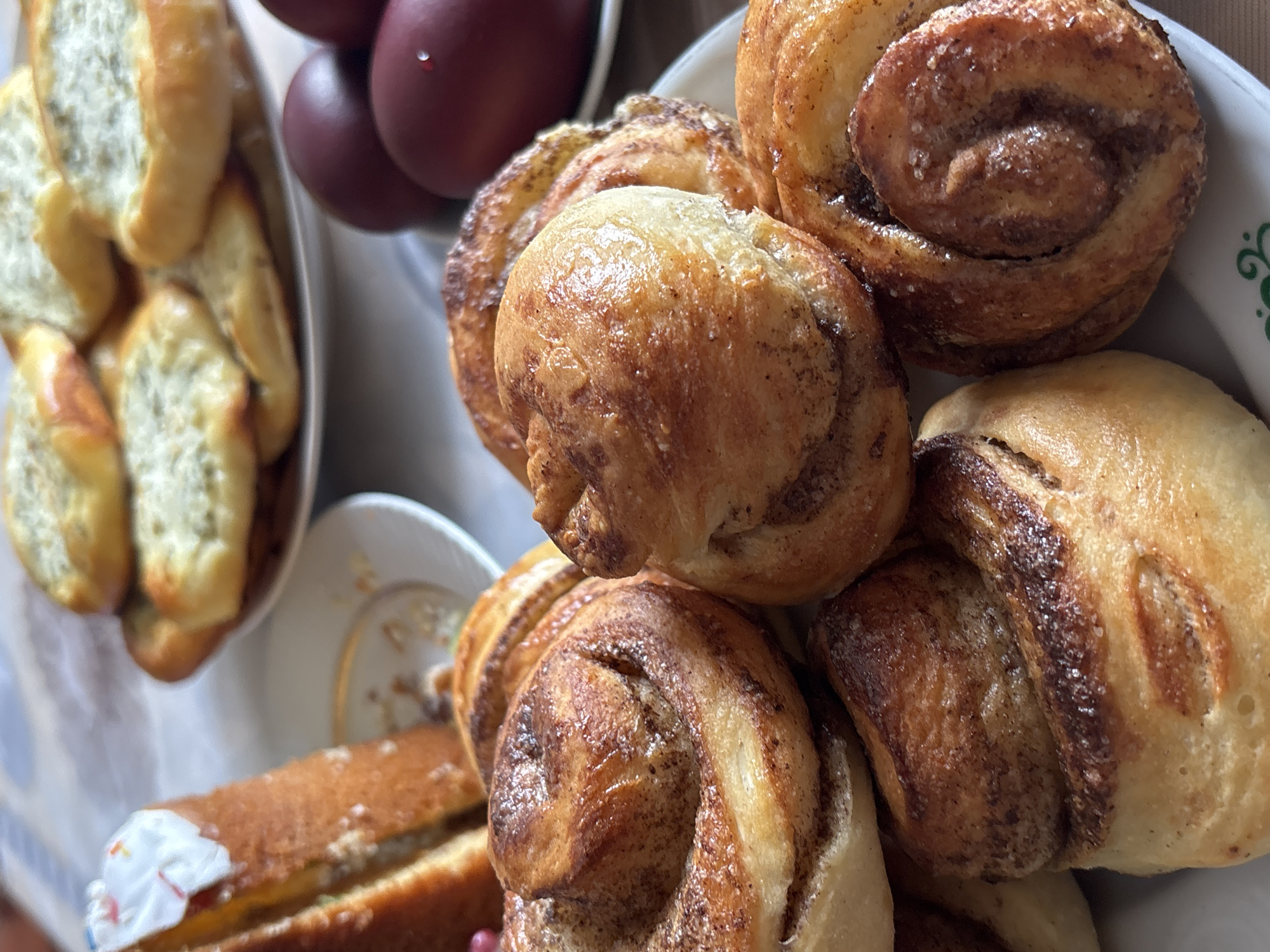 A close-up shot shows an assortment of baked goods arranged on a table. A dish containing small, oblong rolls with a greenish filling is on the left side. Next to it are several deep-red colored eggs. Dominating the foreground is a stack of cinnamon rolls. A cake slice can be seen below the plate with cinnamon rolls. The location is Urada, Russia, but no specific details in the photo provide information about the town's environment or population.