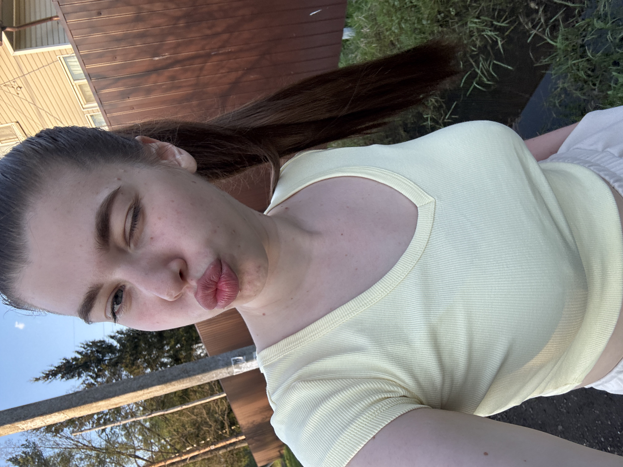 A young woman with brown hair in a high ponytail, wearing a yellow ribbed shirt, is shown in a selfie. She is pouting with her lips pursed. The background includes a wooden building, possibly a house, and green vegetation. The photo appears to be taken outdoors, possibly in Shevlyagino, Russia, given the prompt's instruction. The light suggests it might be late afternoon or early evening.