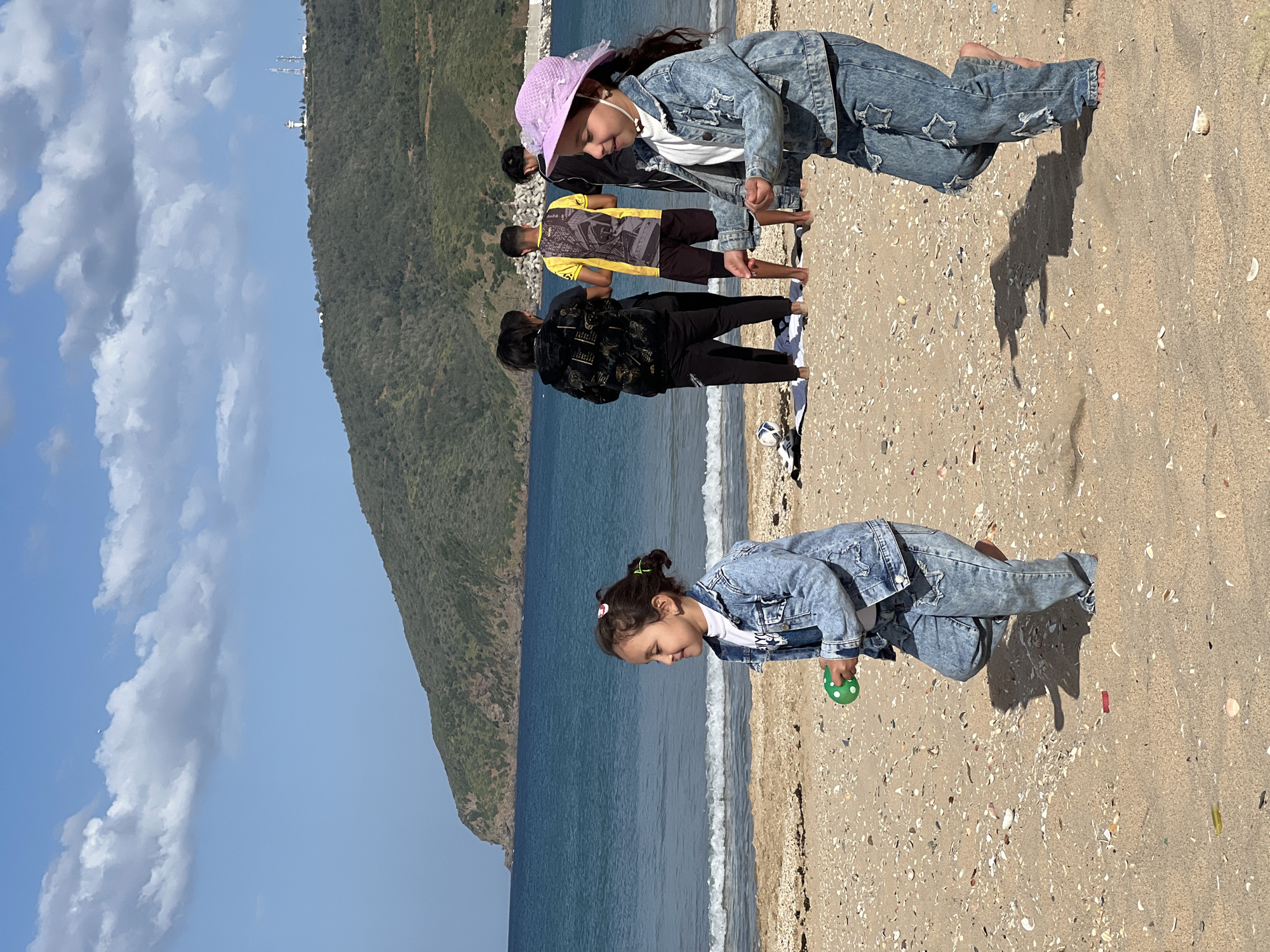 The image shows a beach scene in M'Diq, Morocco. Two young girls wearing similar denim jackets and pants are on the sandy beach. One is wearing a purple hat. Behind them, a group of people stands near the shoreline. A mountain rises in the background. The sky is blue with white clouds. The sea is calm, with gentle waves reaching the shore. The beach is strewn with shells and pebbles. One girl is holding a green, polka-dotted ball.