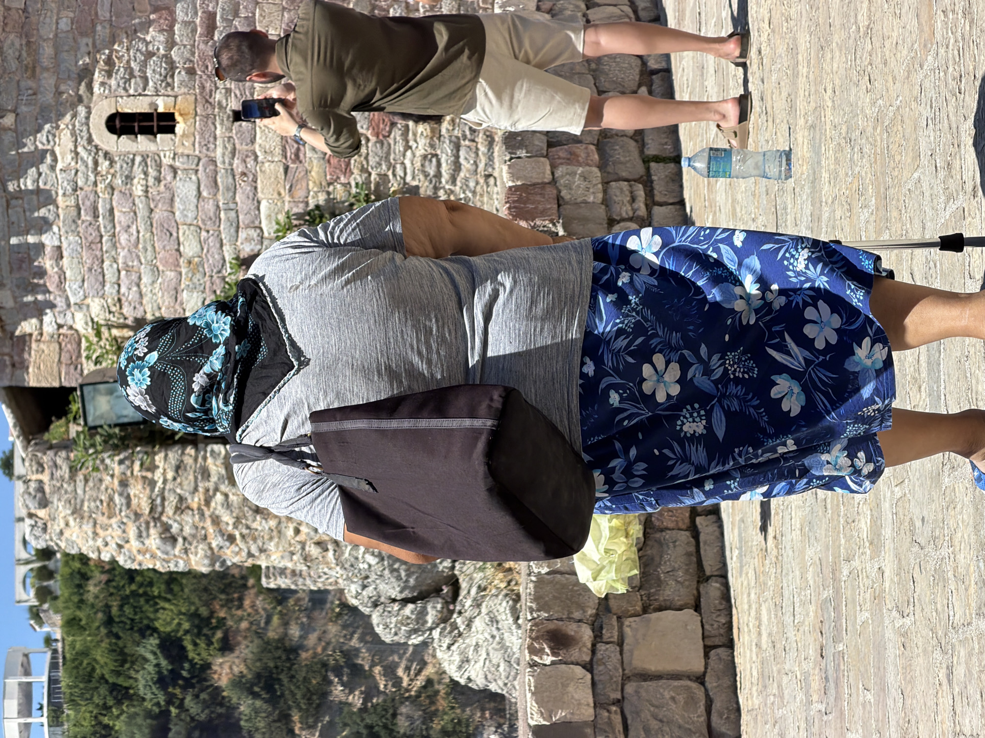 The image captures a scene within the historic Old Town of Budva, Montenegro. In the foreground, a woman wearing a blue floral skirt and a grey t-shirt is seen from behind, with a dark tote bag slung over her shoulder. She appears to be using a walking cane. Her head is covered with a patterned scarf, predominantly black with blue and white floral designs. To her right, a man dressed in a green shirt and beige shorts is taking a photograph with his mobile phone, facing a stone wall with a small, arched window. A plastic bottle of water stands on the cobblestone path between them. In the background, lush green vegetation and structures, including a white building with a rounded roof and arches, are visible on a hillside, suggesting the city's coastal setting and its ancient fortifications. The scene is bathed in bright sunlight, casting shadows on the stone pavement.