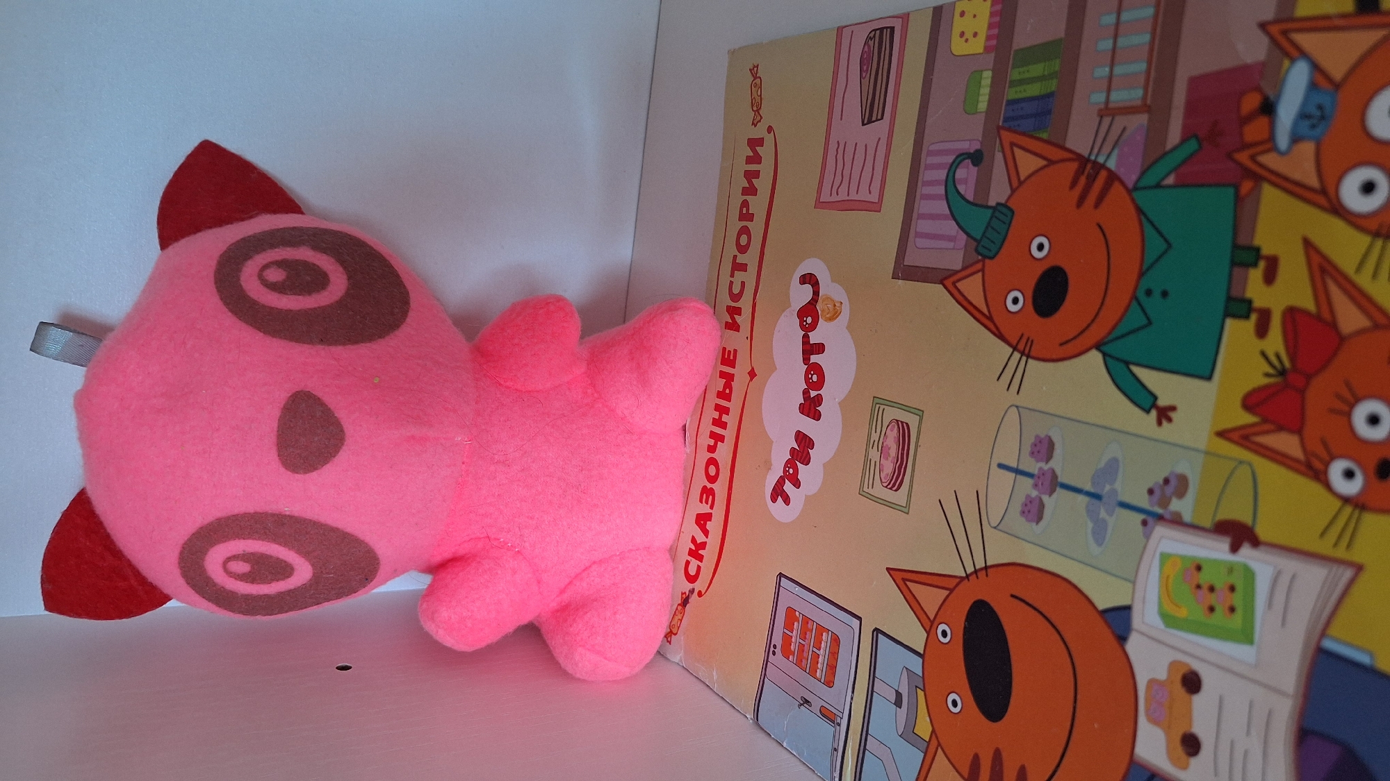 A pink plush toy, possibly a panda, is positioned in the foreground, leaning on a children's book. The book is open, revealing colorful illustrations of cartoon cats and a kitchen scene. The text on the book appears to be in Russian. The background is a plain white surface, likely a shelf or wall. The lighting is bright and even, suggesting an indoor setting. There are no visible people or actions in the image. The image does not contain any specific landmarks or elements that would allow for the determination of its location in Nogliki, Russia.