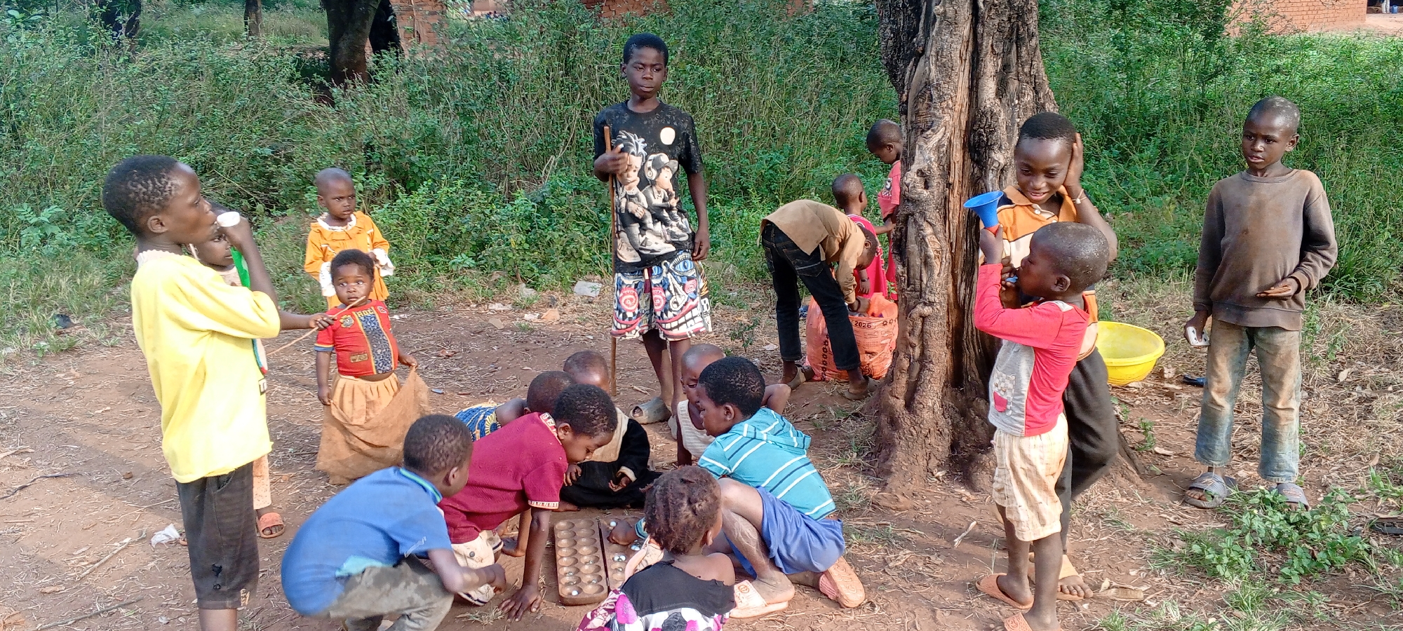 A group of approximately 14 children are gathered in a yard with a large tree. The location is Ngalula, Democratic Republic of the Congo. Some children are playing a board game on the ground. Others stand or lean against the tree, observing. One child is drinking from a white container and another blows into a blue megaphone. A yellow bucket sits near the tree. The ground is dirt and sparse grass. Background vegetation includes bushes and trees.