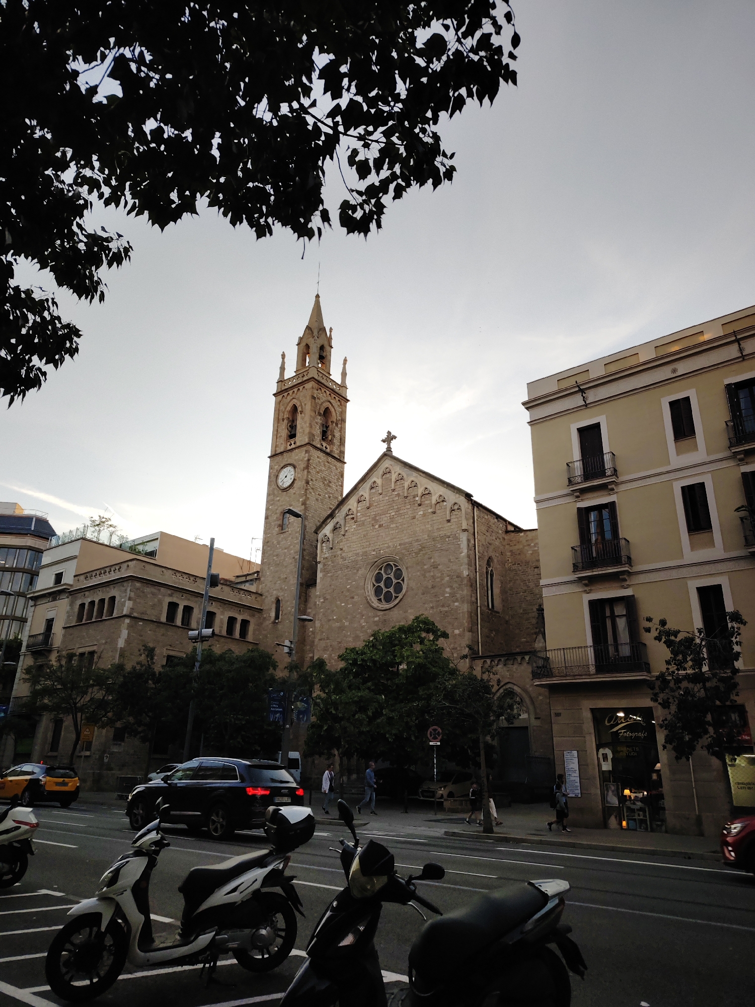 This media file captures an urban street scene in Barcelona, Spain, at dusk or early evening. The sky is a pale, clear grey, indicating the transition from day to night.
The central subject is a historic stone church featuring a prominent bell tower with a spire and a visible clock face. The main facade of the church also displays a large, circular rose window and is topped by a cross. To the right of the church stands a multi-story building with a light yellow facade, dark window frames, and balconies. To the left, another building is visible with what appears to be rooftop greenery.
In the foreground, several motorcycles and scooters, including white and dark models, are parked along the street. The road hosts various vehicles, such as a yellow taxi, a dark SUV (with license plate "0626 LKF"), and a partially visible red car. Pedestrians can be seen walking on the sidewalks, adding to the typical urban activity. Silhouetted tree branches and leaves frame the top of the image.
Notable details include a shop sign on a building to the right of the church, which reads "Artes Fotografia." The number "301" is visible above this shop. A white circular sign with a red border and diagonal line, indicating a "no parking" or "no stopping" restriction, is also present on the sidewalk. The architecture, including the church's design and the style of the surrounding buildings, is characteristic of European cities like Barcelona.