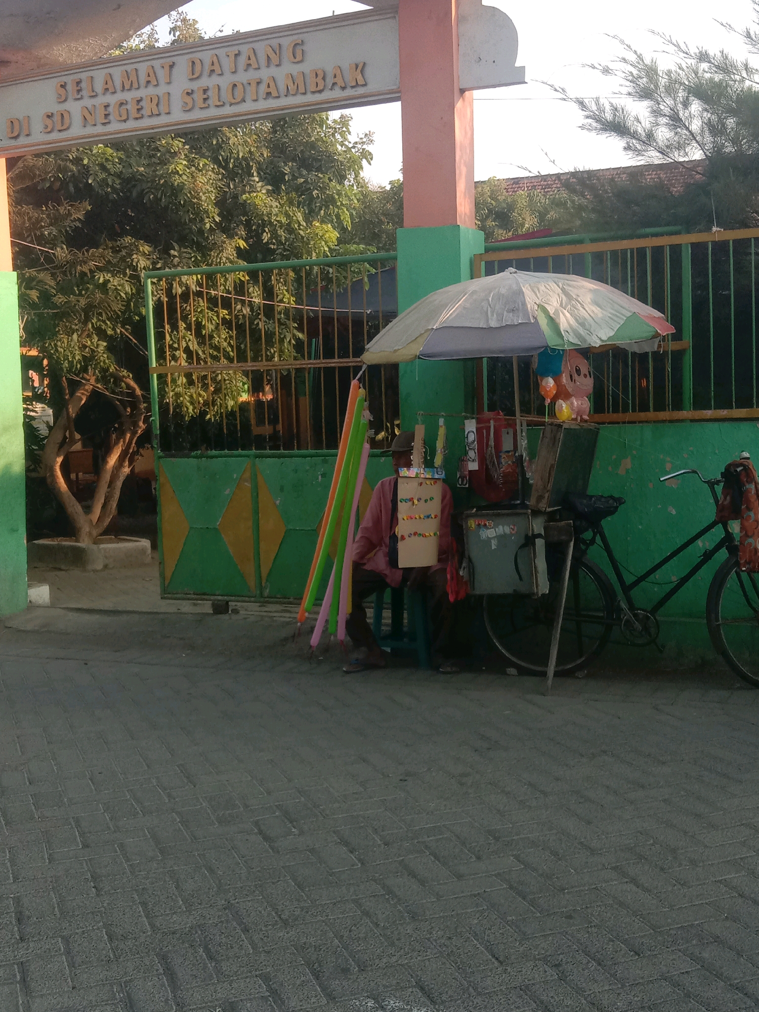 This image depicts the entrance of a school in Selotambak, Indonesia, identified by the signage "SELAMAT DATANG DI SD NEGERI SELOTAMBAK". A vendor is seated under an umbrella, attending to a cart filled with various items, including balloons. Beside the cart, a bicycle is parked, with a patterned cloth draped over its handlebars. The ground is paved with interlocking bricks. The scene appears to be set outdoors, possibly in the late afternoon, given the lighting and shadows.