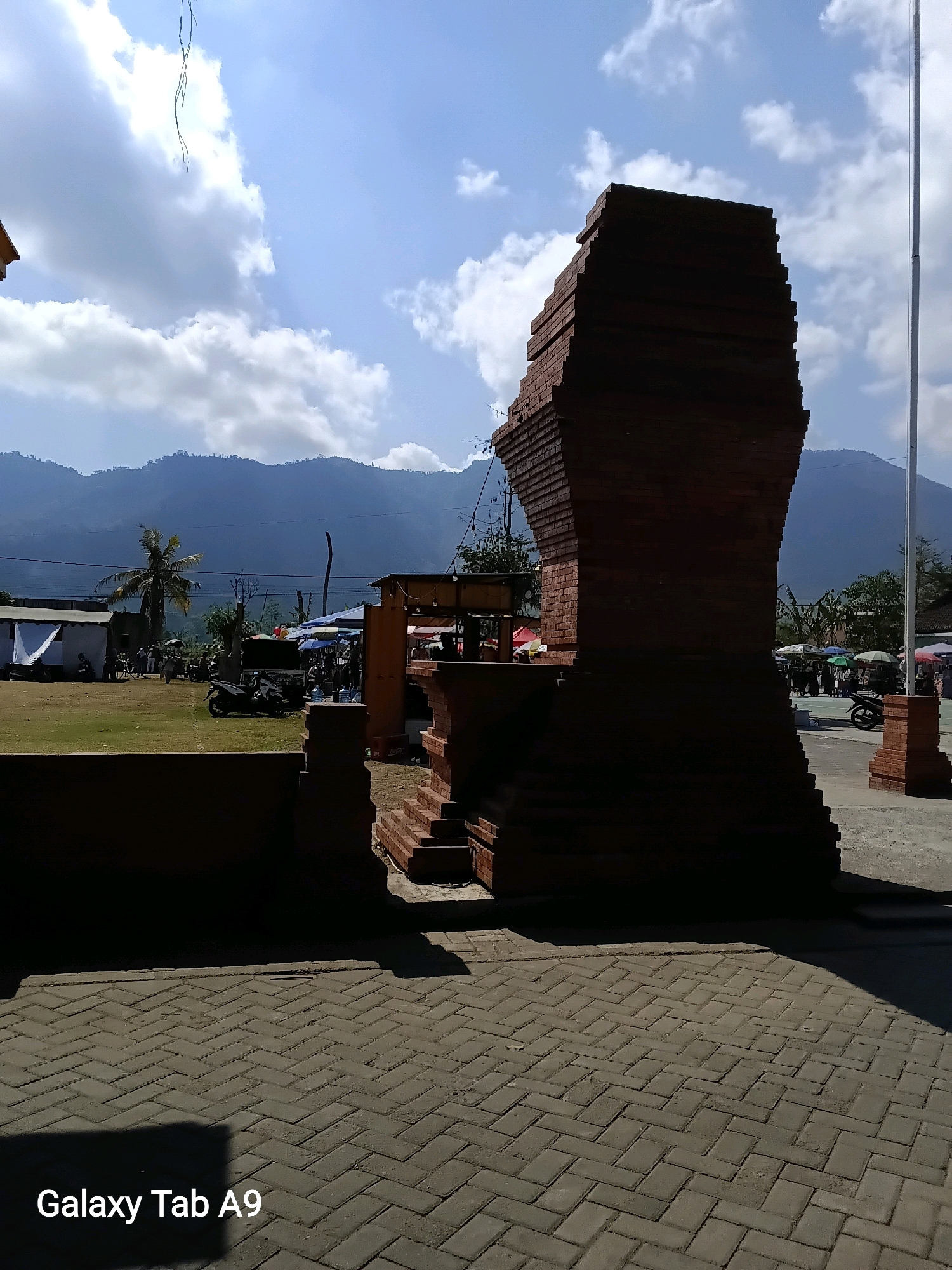 The image captures a sunny day in Ngadirenggo, Indonesia, with a prominent brick structure in the foreground, possibly an archway or monument. The structure is dark and silhouetted against a bright sky with scattered white clouds. Behind the brick structure, a grassy field is visible with several small buildings and tents, suggesting a local market or event. In the background, a line of lush green mountains forms a scenic backdrop under the clear blue sky. Several motorcycles are parked in the field, and a few people can be seen walking around the tents. A tall flagpole stands to the right of the brick structure. The foreground is paved with gray interlocking bricks laid in a herringbone pattern, with a shadow cast across it from the brick structure. A watermark at the bottom left corner reads "Galaxy Tab A9".