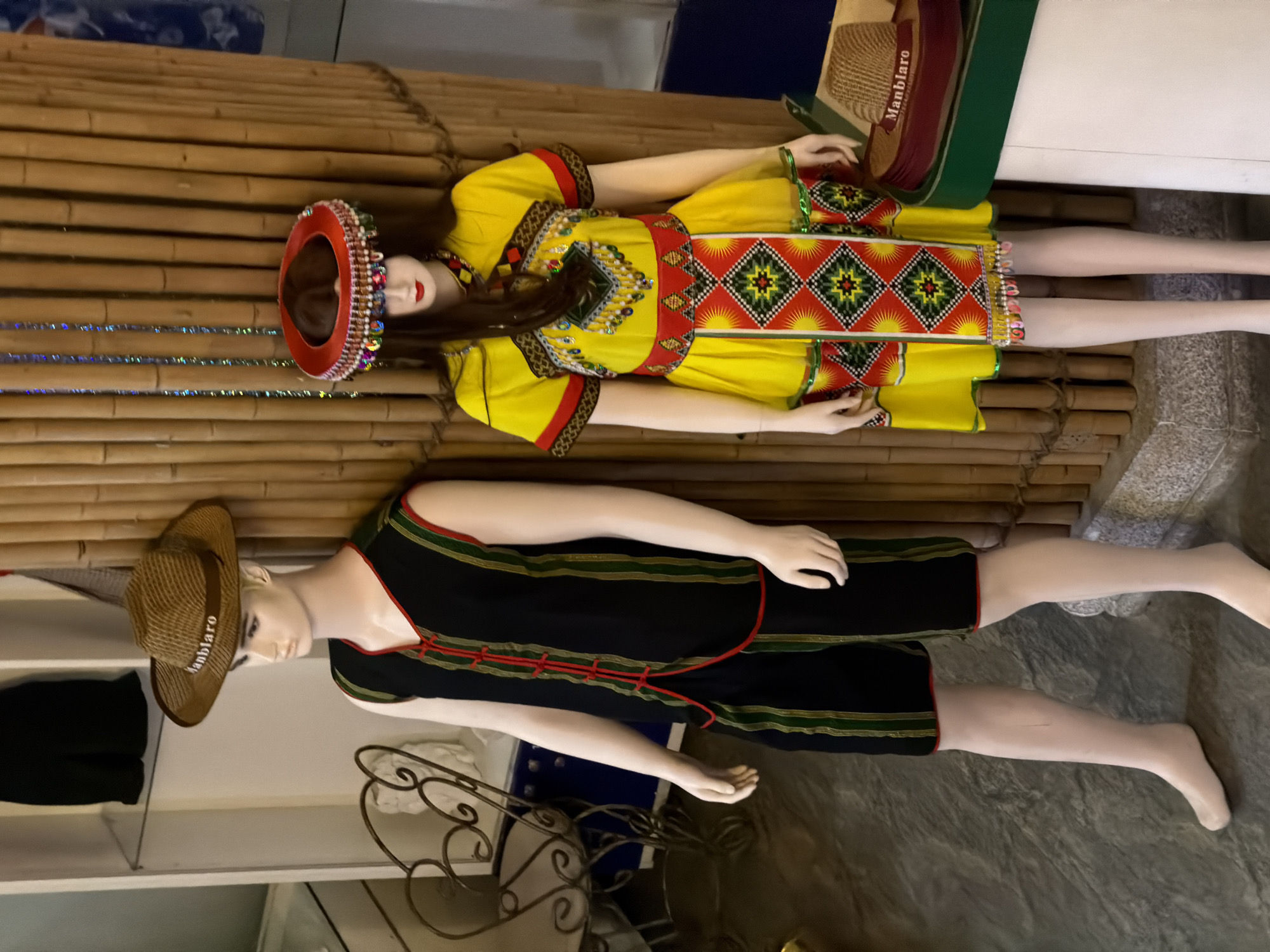 In Nam Nha Trang Ward, Vietnam, two mannequins are displayed against a backdrop of vertical bamboo poles. The mannequin on the left, appearing male, is wearing a woven straw hat with the word "Manblaro" printed on a band, a sleeveless black vest with red ties and green and gold stripes, and matching black shorts with similar striped detailing. The mannequin on the right, appearing female, is adorned in a bright yellow dress with colorful geometric patterns and intricate beadwork around the neckline. She also wears a red headdress embellished with beads and sequins. A woven straw hat with a burgundy band that reads "Manblaro" is placed on a green surface to the right of the female mannequin, atop a white cylindrical structure. The floor is a dark, textured stone.