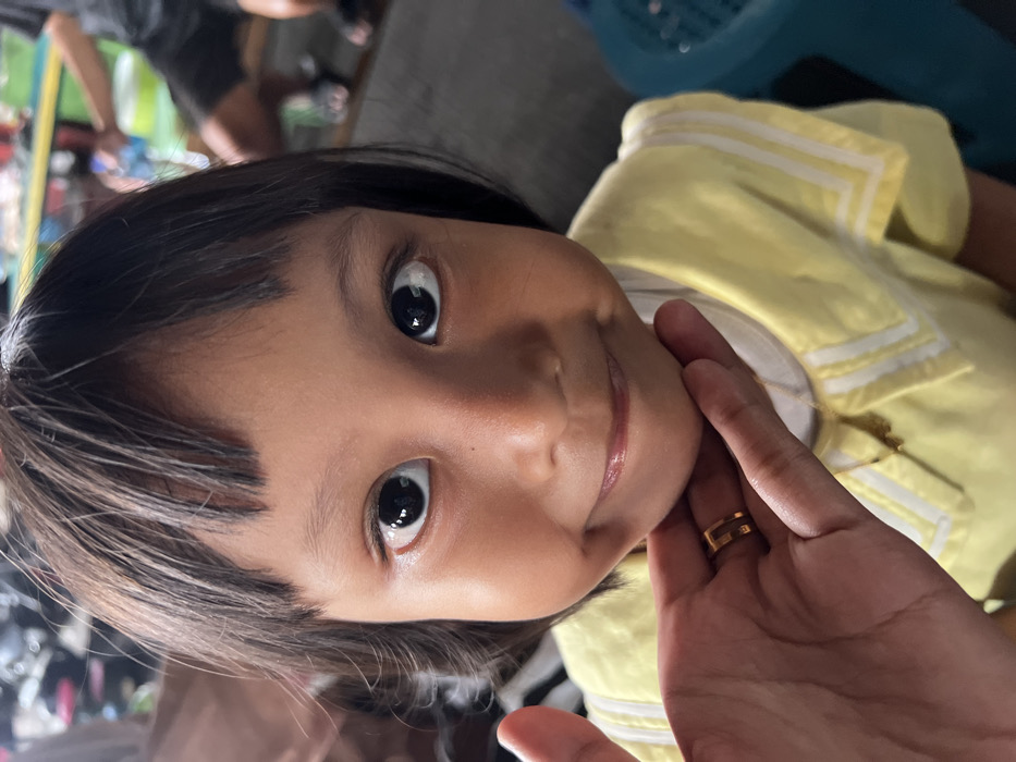 The image shows a close-up portrait of a young child, likely female, in Nganguk, Indonesia. The child has dark hair with straight bangs and a bob cut, wide dark eyes, and a subtle closed-mouth smile with full cheeks. She wears a light yellow top featuring a white collar with parallel white stripes, and a faint gold-colored necklace. An adult's hand, displaying a medium skin tone and a gold ring with a dark horizontal band on the ring finger, is gently cupping the child's chin from below. The background is out of focus but indicates an indoor or covered outdoor public setting. Visible elements include indistinct shapes resembling parked motorbikes on the upper left, and a person's lower body clad in dark shorts and sandals, alongside a blue plastic object, on the right.