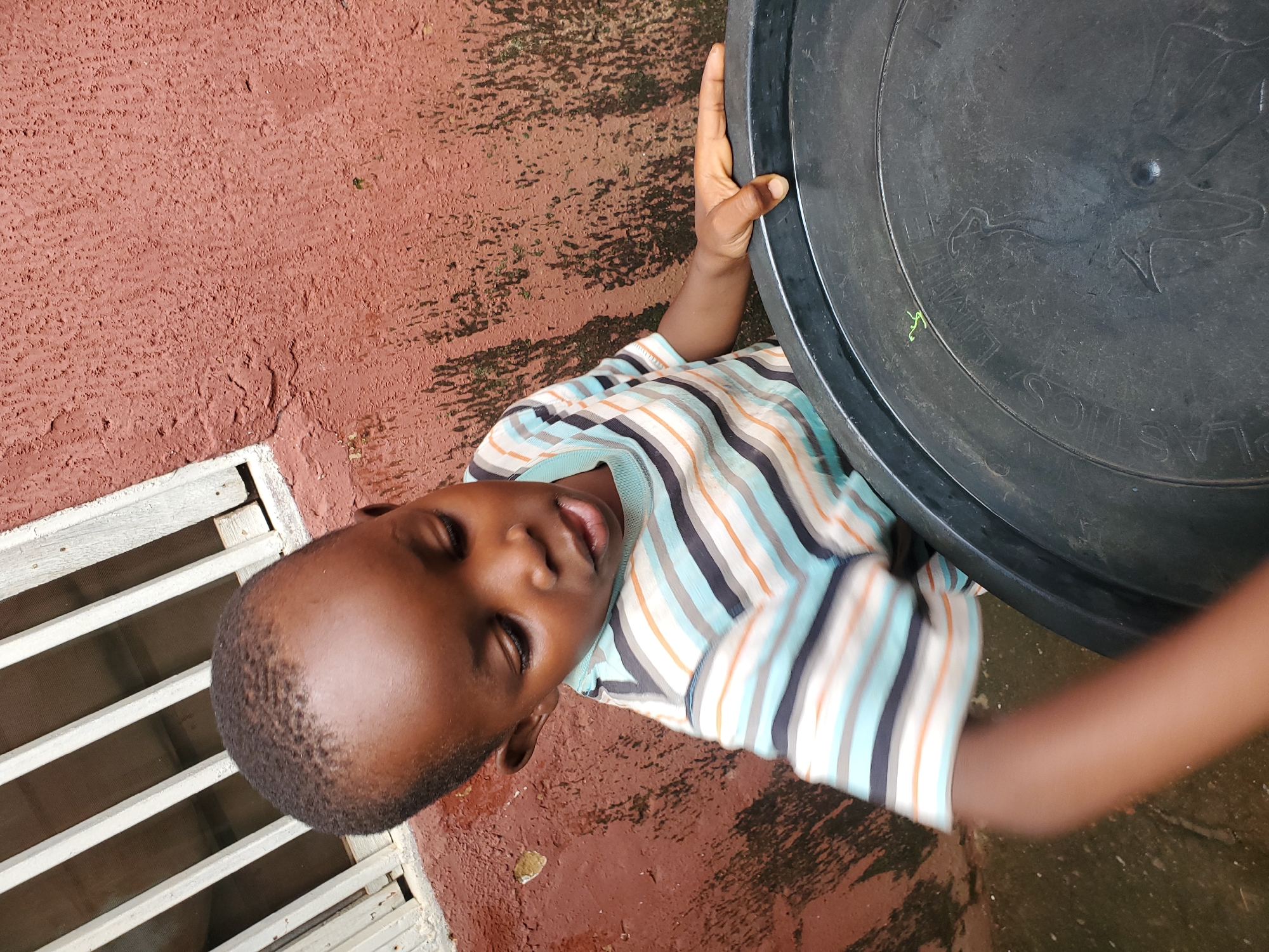 This is a portrait-oriented, medium shot of a young boy in an outdoor setting. The boy, who appears to be of African descent, is wearing a horizontally striped t-shirt in shades of blue, white, and orange. He is looking upwards with his eyes closed and his mouth slightly open, suggesting a moment of rest or perhaps an expression of emotion. His skin tone is dark brown, and his hair is short and dark.
To the boy's right, a large, black plastic container dominates the right side of the frame. The container has some embossed text and imagery, though much of it is obscured or difficult to read. One visible word appears to be "PLASTICS". The boy's right arm is extended, and his hand is resting on the rim of this container.
The background consists of a textured, reddish-brown surface that appears to be a wall or ground, possibly damp or wet in places, with some dark shadows cast upon it. To the boy's left, there is a white grating or ventilation system, suggesting the setting might be near a building. The lighting indicates it is daytime, with the sun casting shadows. The overall impression is one of an informal, possibly humid environment. The image has been rotated so that the subjects appear to be lying down, rather than standing.