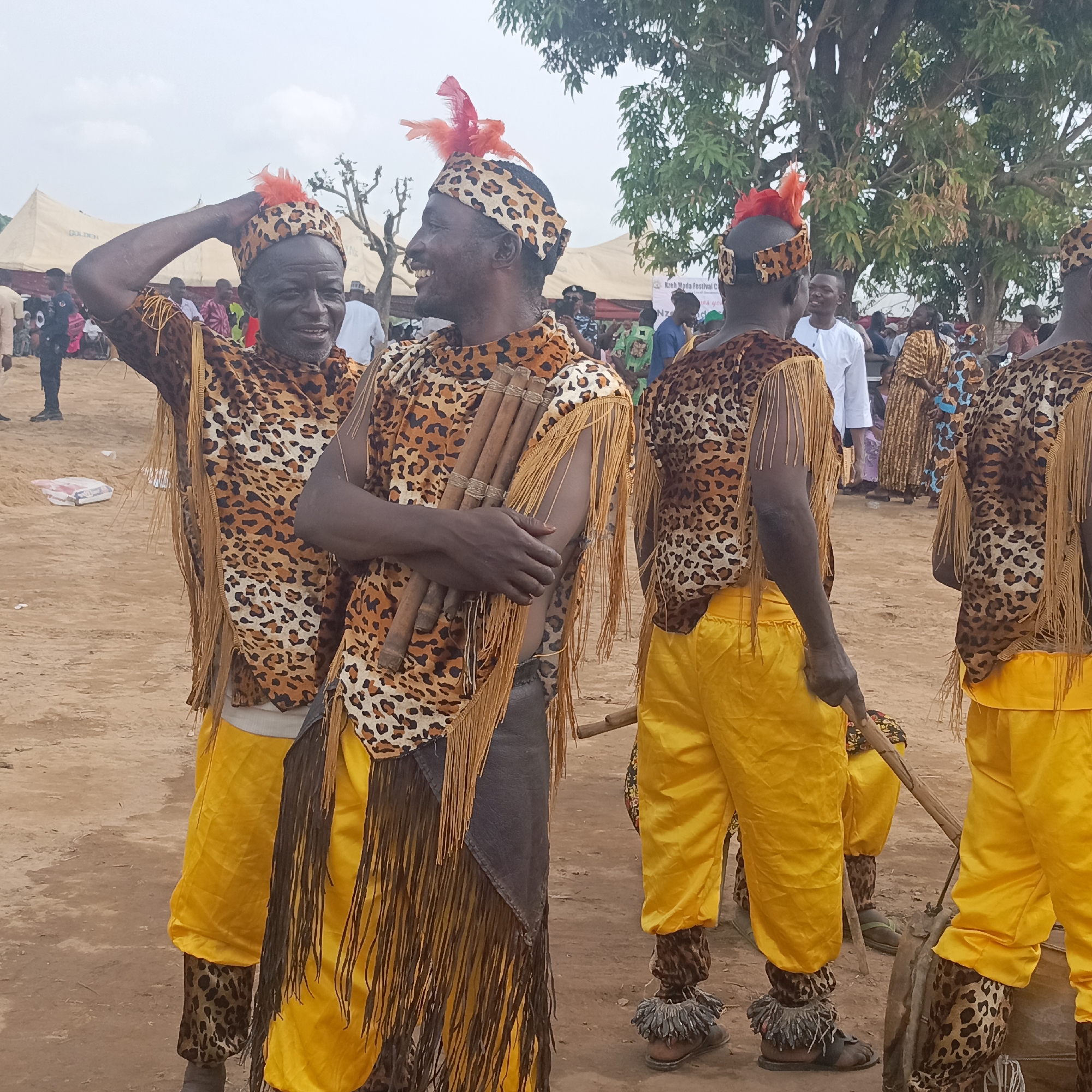 The image shows a group of men, likely performers, at an outdoor event in Akwanga, Nigeria. The men are dressed in traditional attire including leopard-print tops and headwear with orange feathers, yellow pants, and fringe detailing. The man in the center holds a set of wooden pipes or instruments. The background shows a crowd of people, tents, and trees. The ground is dirt or sand.