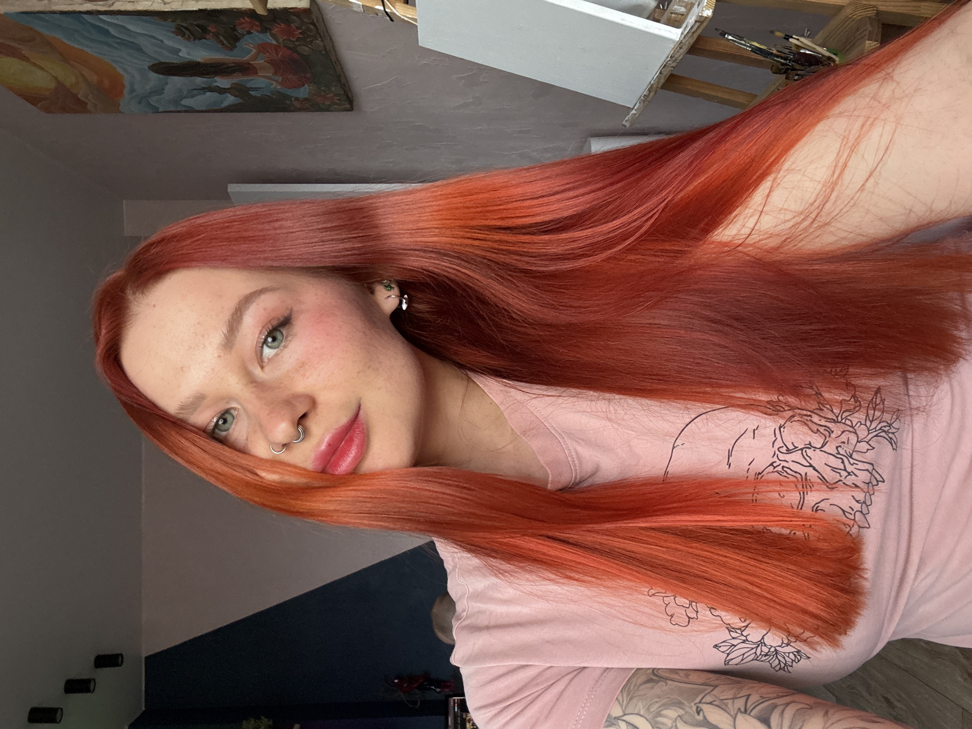 A portrait of a young woman with vibrant red hair taking a selfie indoors. She has fair skin with freckles, green eyes, a nose ring, and a pink t-shirt with a floral design. Her hair is long and flowing. In the background, there is a painting on the wall, an easel, and paintbrushes. The lighting is soft and diffused. Based on the background, the location might be an artist's studio or a home with artistic elements. There's no definitive evidence to confirm the specific location of Rostov-on-Don, Russia; that information is not present in the image.