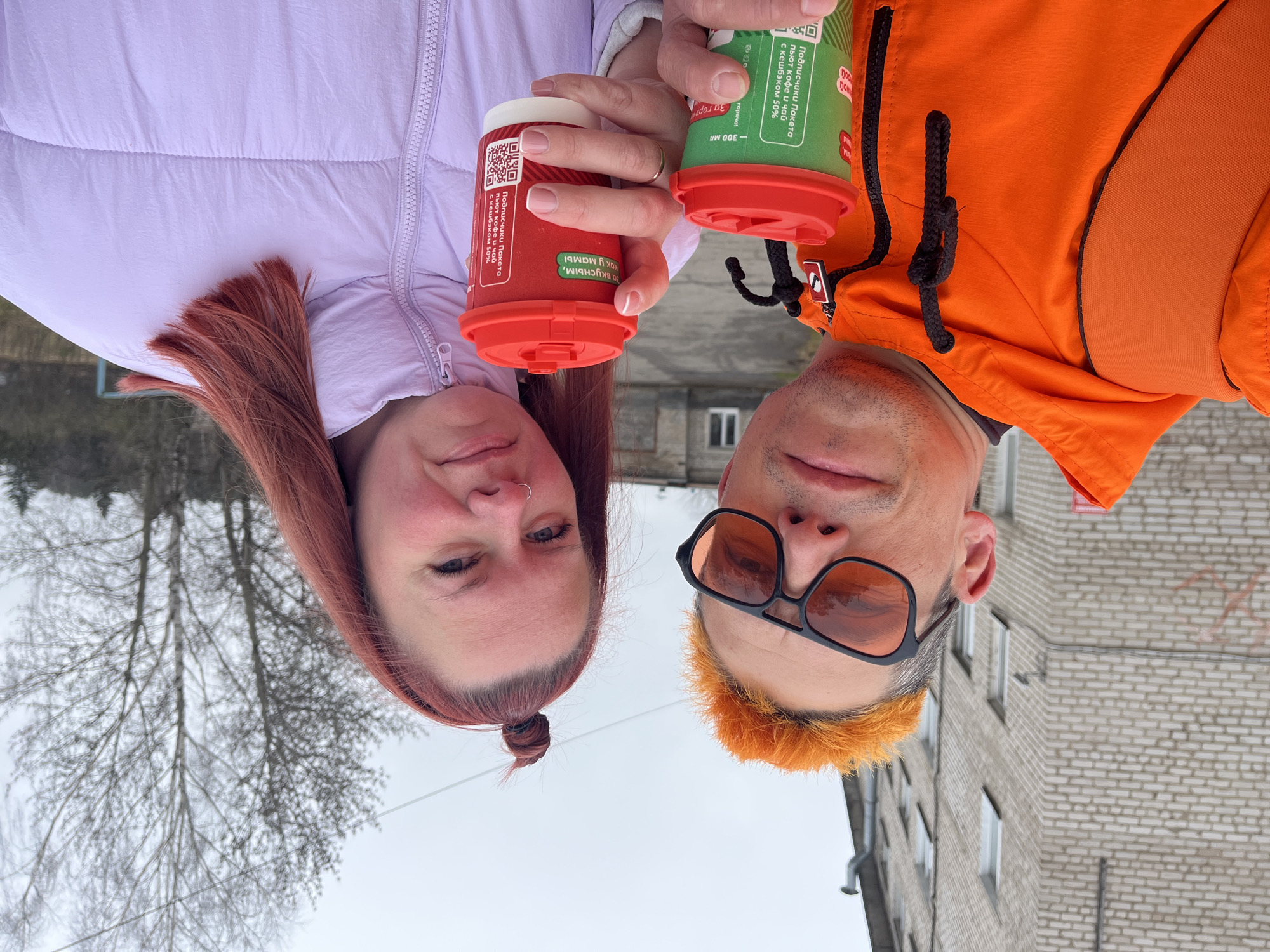 The image shows two individuals, a woman and a man, holding takeaway coffee cups. They appear to be posing for a selfie, with their faces positioned close together. The image is oriented upside down.
The woman has red hair, a piercing on her left nostril, and is wearing a light purple puffer jacket. The man is wearing orange sunglasses and an orange hooded jacket. His hair is dyed orange. They are each holding a coffee cup; hers is red, and his is green. Both cups have red lids.
The background reveals an urban setting with buildings constructed from light grey brick. Trees with bare branches are visible in the distance. There is text in Russian on the cups. Based on context clues from Russian-language social media posts using the same image, the location can be inferred to be in the city of Chaikovsky, Perm Krai, Russia.