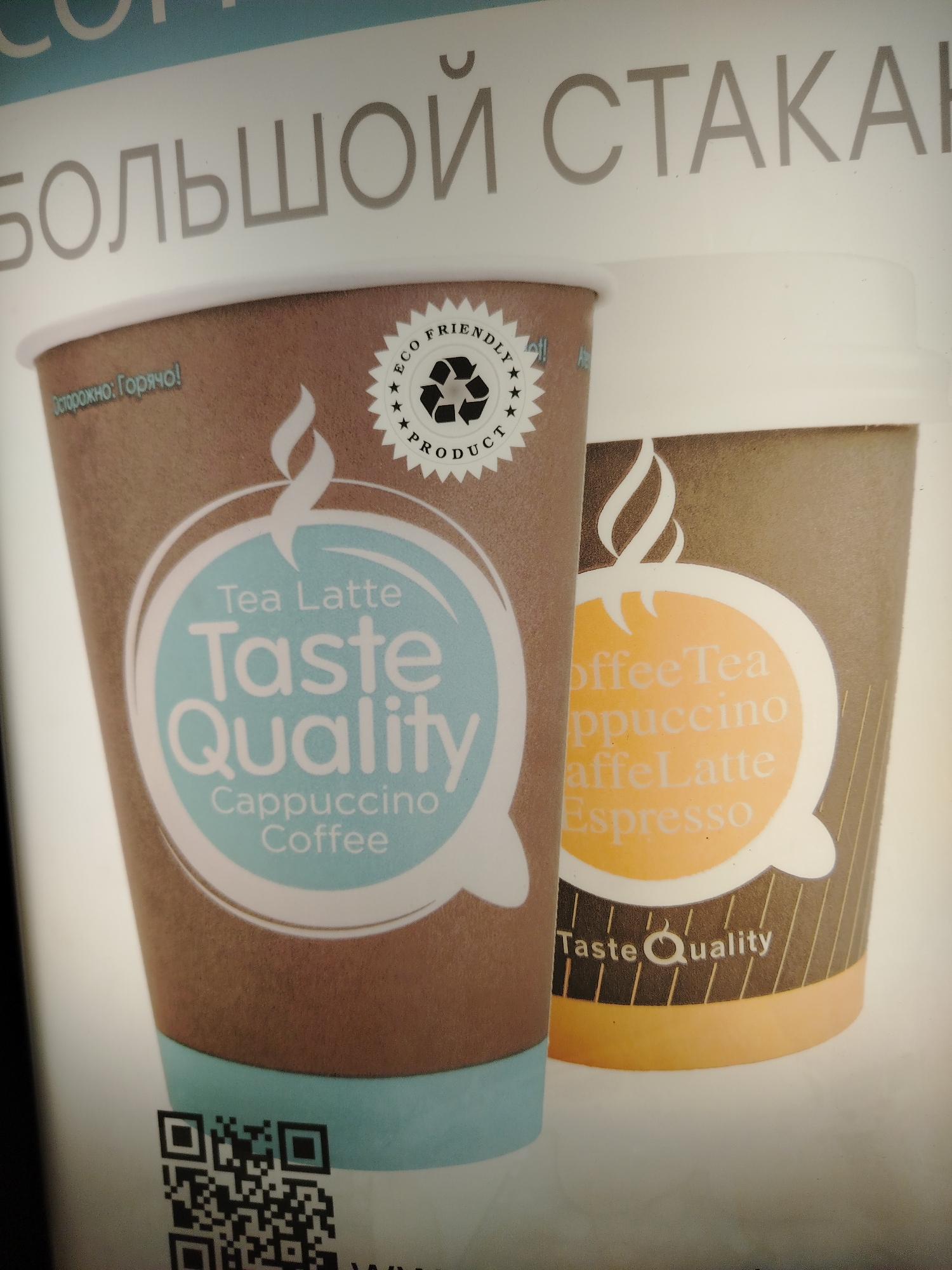 An upward-angled shot captures an advertisement or display featuring two disposable beverage cups, likely for a vending machine or cafe. The foreground cup, predominantly brown with a light blue bottom rim, features a white steam icon above a circular label. This label, with a light blue background, reads "Tea Latte," "Taste Quality," "Cappuccino," and "Coffee" in white text. Above it, the Russian text "Осторожно: Горячо!" (Caution: Hot!) is visible. A star-shaped badge with "ECO FRIENDLY" and "PRODUCT" encircles a recycling symbol on the upper right of this cup.
Behind it, a second cup, partially obscured, shows a brown and orange design. Its orange circular label displays partially visible text like "offee Tea," "ppuccino," "affeLatte," and "Espresso." Below this label, the words "Taste Quality" are also visible. In the bottom left of the foreground cup, a black and white QR code is present. At the top of the image, large Russian text reads "БОЛЬШОЙ СТАКА" (part of "БОЛЬШОЙ СТАКАН" meaning "BIG CUP") against a light background. The overall scene is brightly lit, focusing on the products advertised in Rusanovo, Russia.